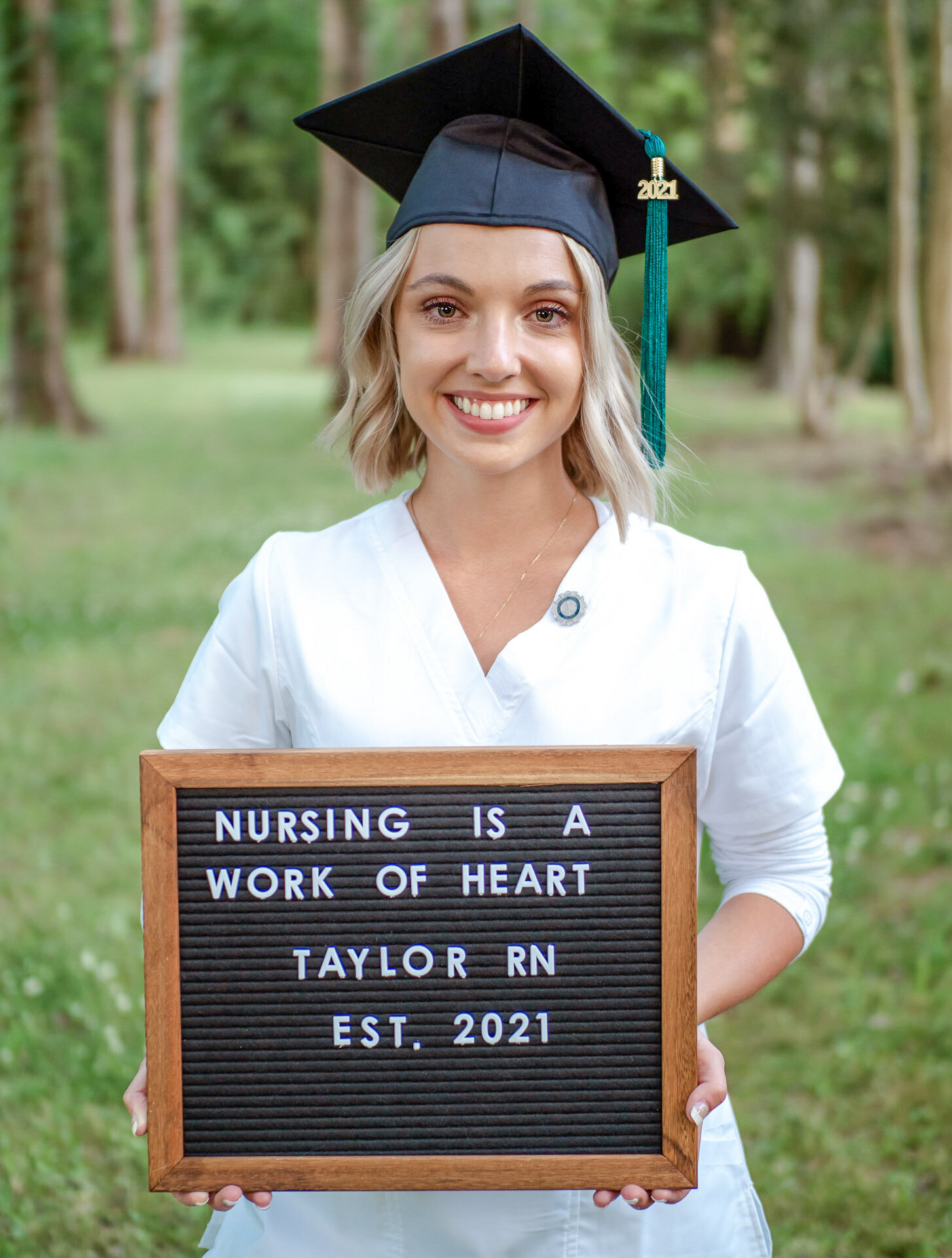 A UNCW nursing graduate in a cap and gown holding a message board that reads "Nursing is a Work of Heart" in a wooded area in Wilmington, North Carolina. This portrait captures academic achievement and personal dedication, perfect for celebrating graduation.