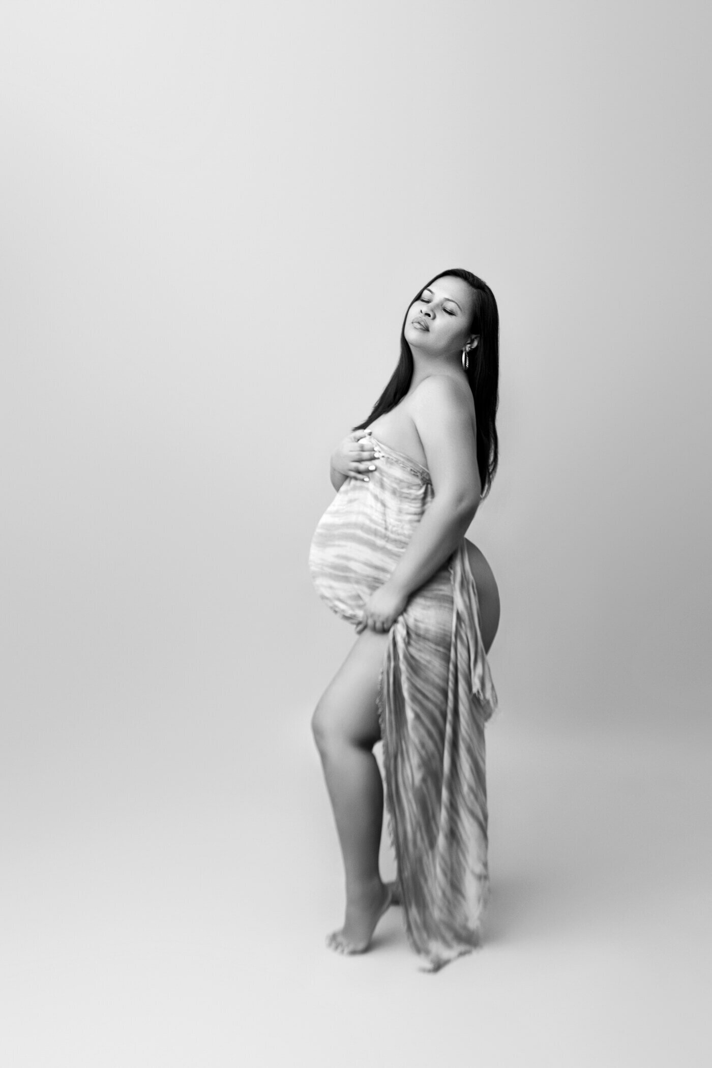 A black-and-white portrait of a pregnant woman standing on a plain background, wrapped in a flowing, striped fabric that drapes elegantly around her body. She cradles her baby bump with one hand and holds the fabric with the other, exuding confidence and serenity. Her eyes are closed, and she has a peaceful expression, capturing the beauty and strength of motherhood.