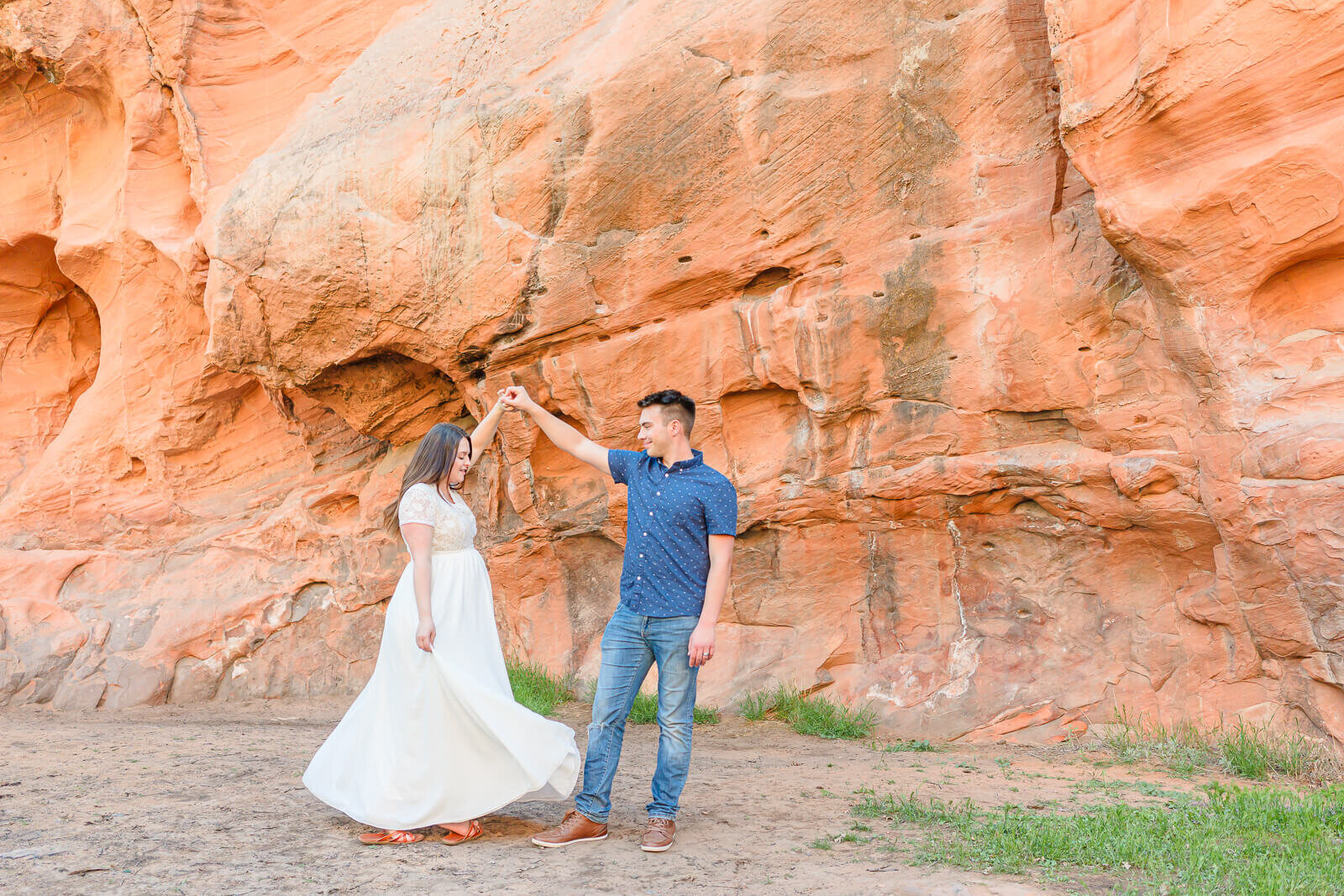 A man wearing a blue button-down shirt and blue jeans twirls his pregnant wife wearing a long white maternity dress. Captured at Red Ledges in Spanish Fork Canyon