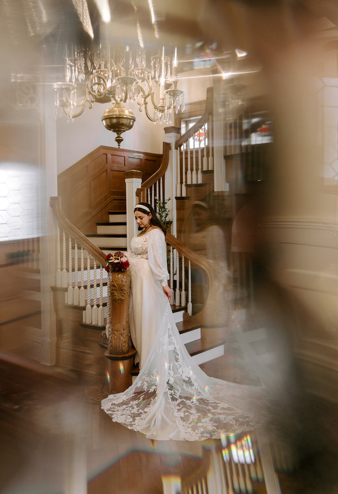 Bride in elegant gown stands on a wooden staircase, her look enhanced by modern bridal makeup trends. A chandelier glows above while a bouquet rests on a nearby pillar, completing the enchanting scene.