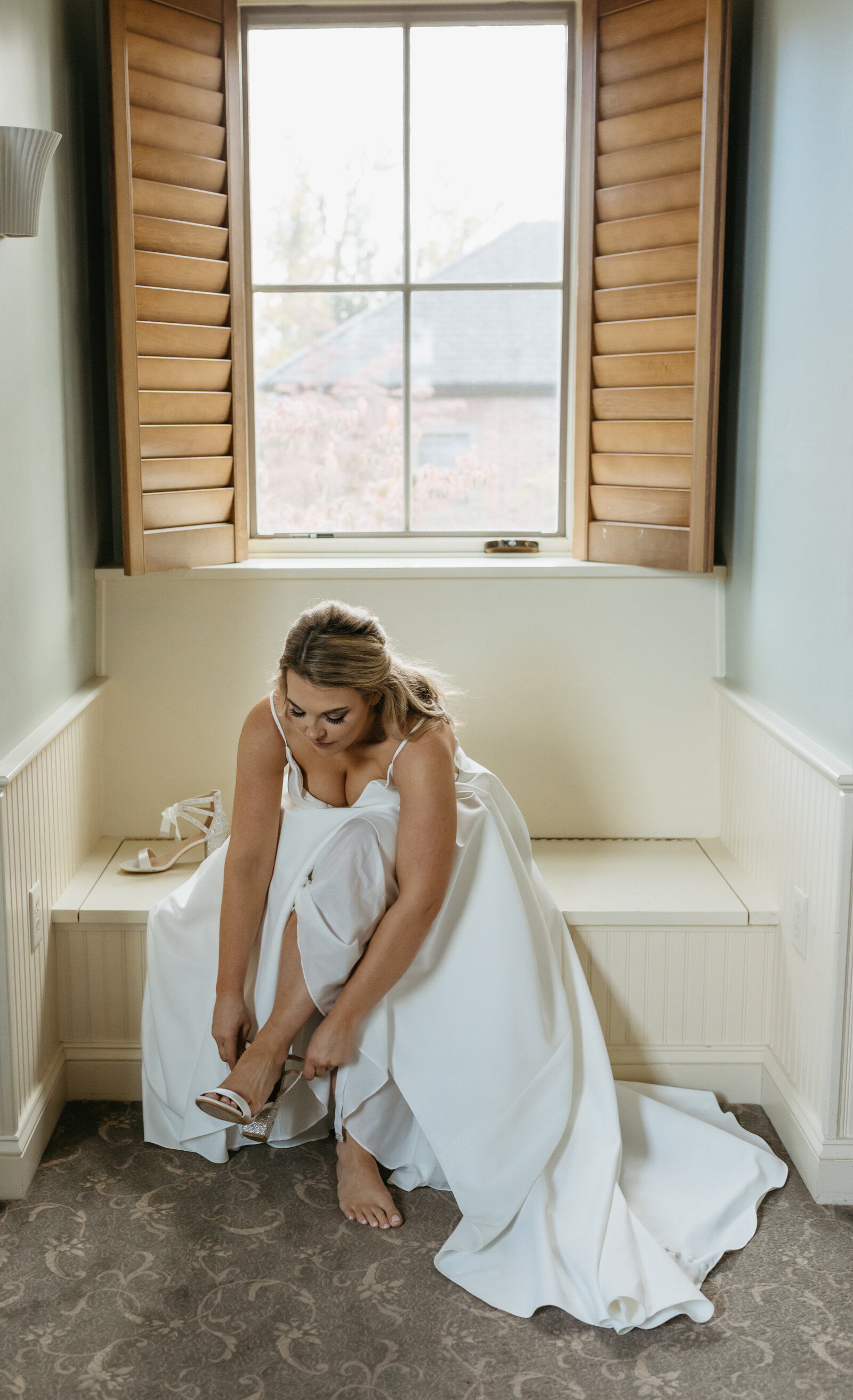 A bride sitting in a window seat fastens her shoes