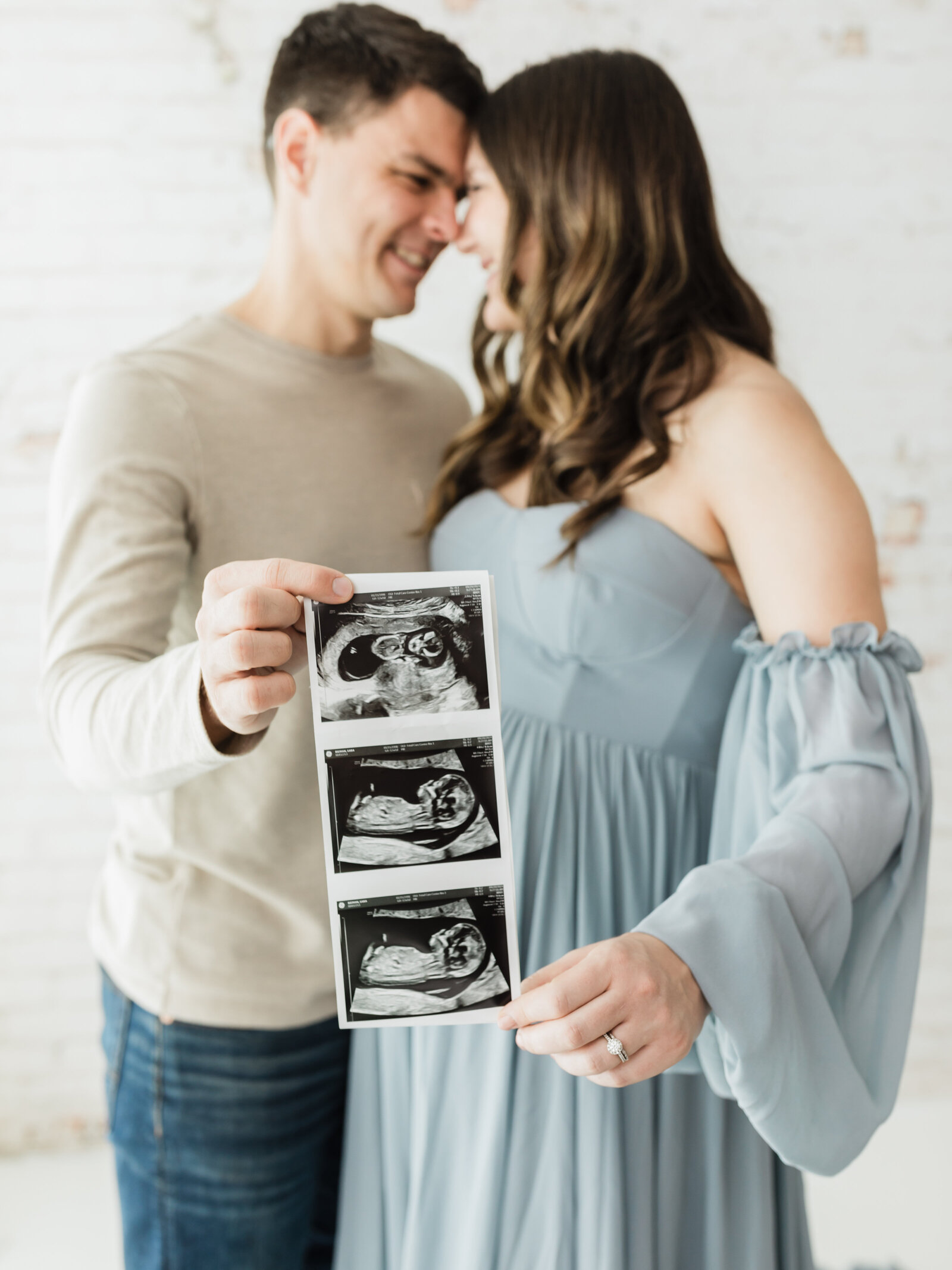 husband and pregnant wearing blue maternity gown holding up ultrasound photo for maternity studio photoshoot