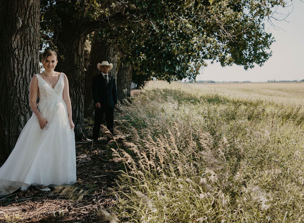 Bride and groom posing in trees beside a scenic field at Bergthal Event Centre, offering a picturesque outdoor wedding setting in Alberta