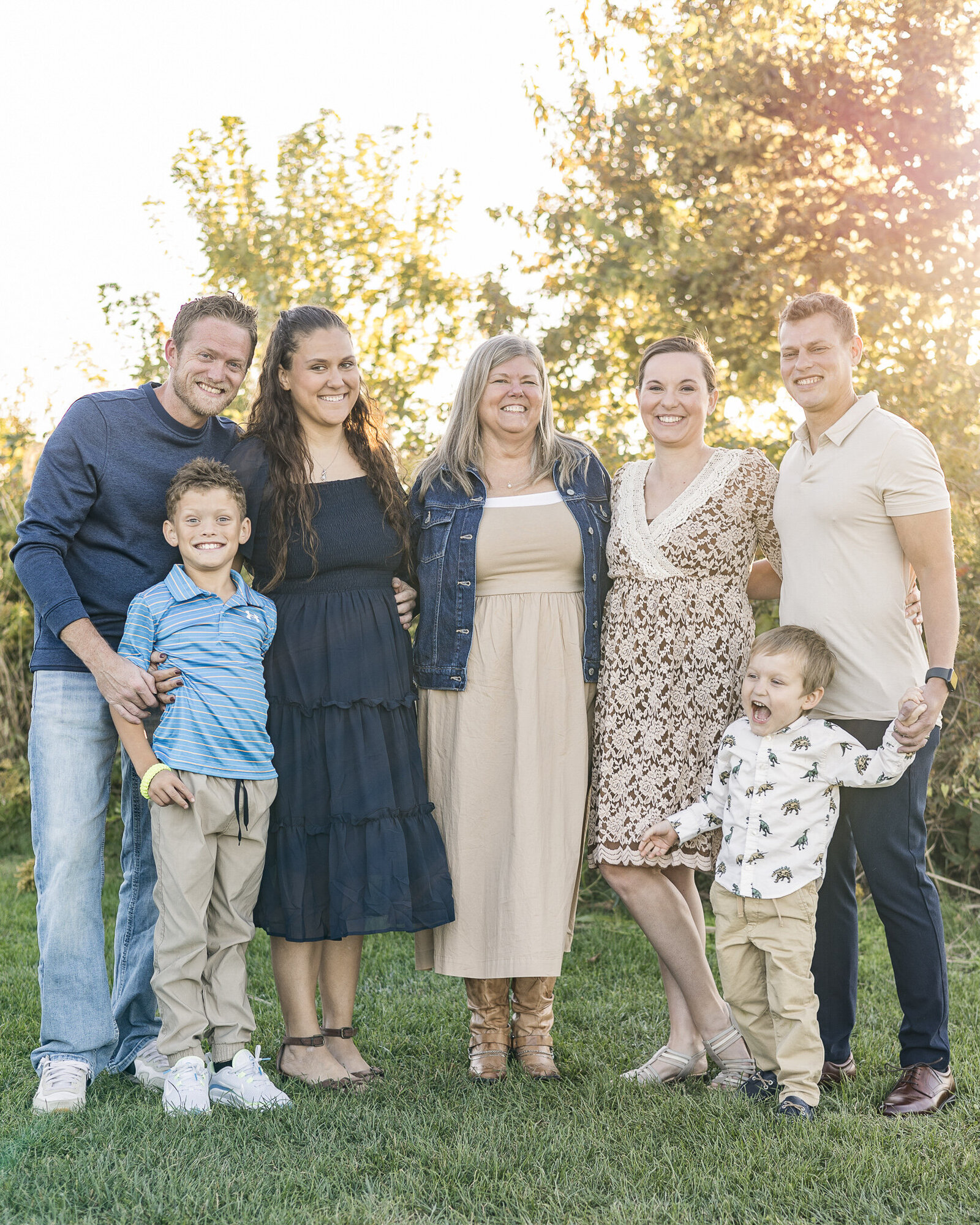 An extended family session with grandma and her son and daughter with their family.  They are standing on the grass in a park.  Grandma is in the middle wearing a blue jean jacket, tan dress, and tan boots.  One son's family is standing at one side.  Dad has sandy blonde hair, is wearing a dark blue long sleeve shirt smiling at the camera.  Mom has dark brown hair, is wearing a dark blue dress smiling at the camera.  Their son is standing in front of them, has sandy blonde hair, smiling at the camera, wearing a light blue and white striped shirt.  Her other son and family is standing on her other side.  Dad has dark brown hair, wearing a light tan short, dark blue jeans, smiling at the camera.  His wife is wearing a tan and white dress with dark brown hair, smiling at the camera.  Their young son is wearing a white print shirt with khaki pants.