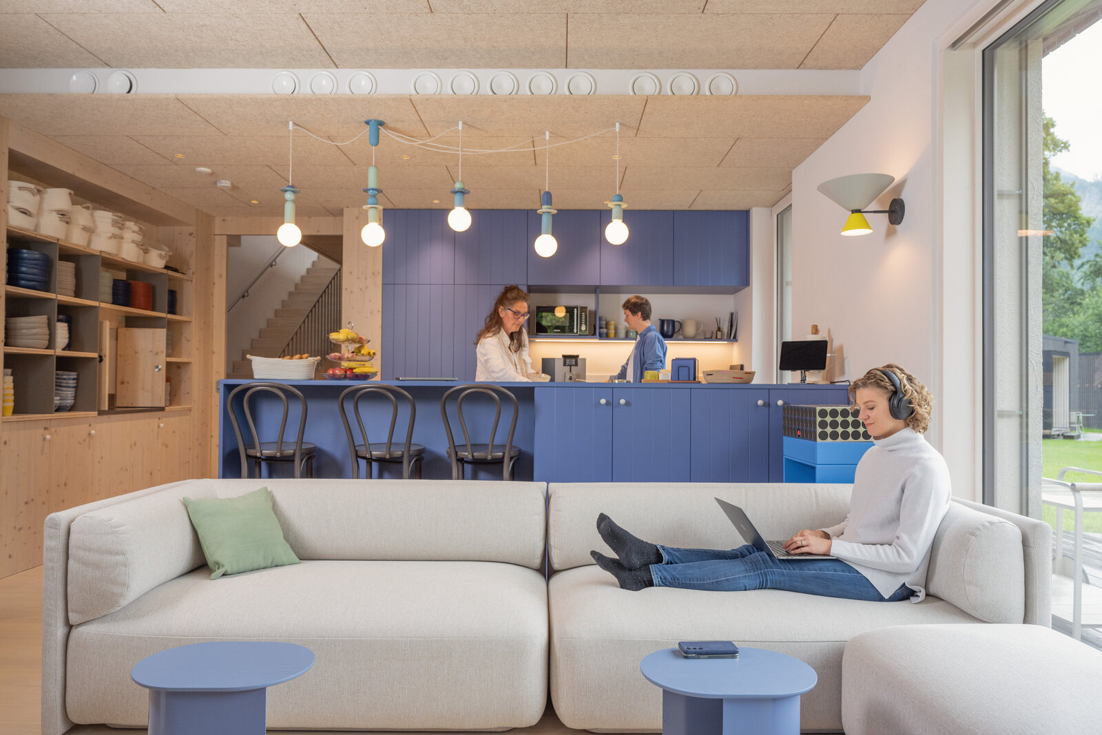 Woman working on a couch with her laptop, while colleagues make coffee in the kitchen background at The Campus Alps Workbase, providing a relaxed and collaborative atmosphere for remote work and company retreats in the Alps.
