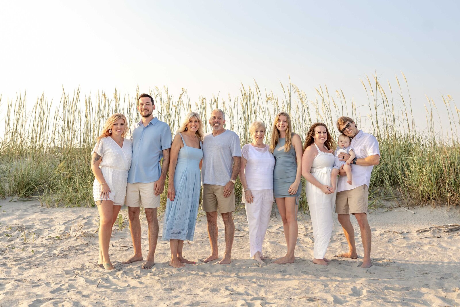 A family poses on sandy dunes, surrounded by swaying sea oats and dressed in pastel and white outfits. The bright sky adds warmth to the scene, perfect for artistic family photography in Wilmington, NC, capturing natural beauty and family connections.