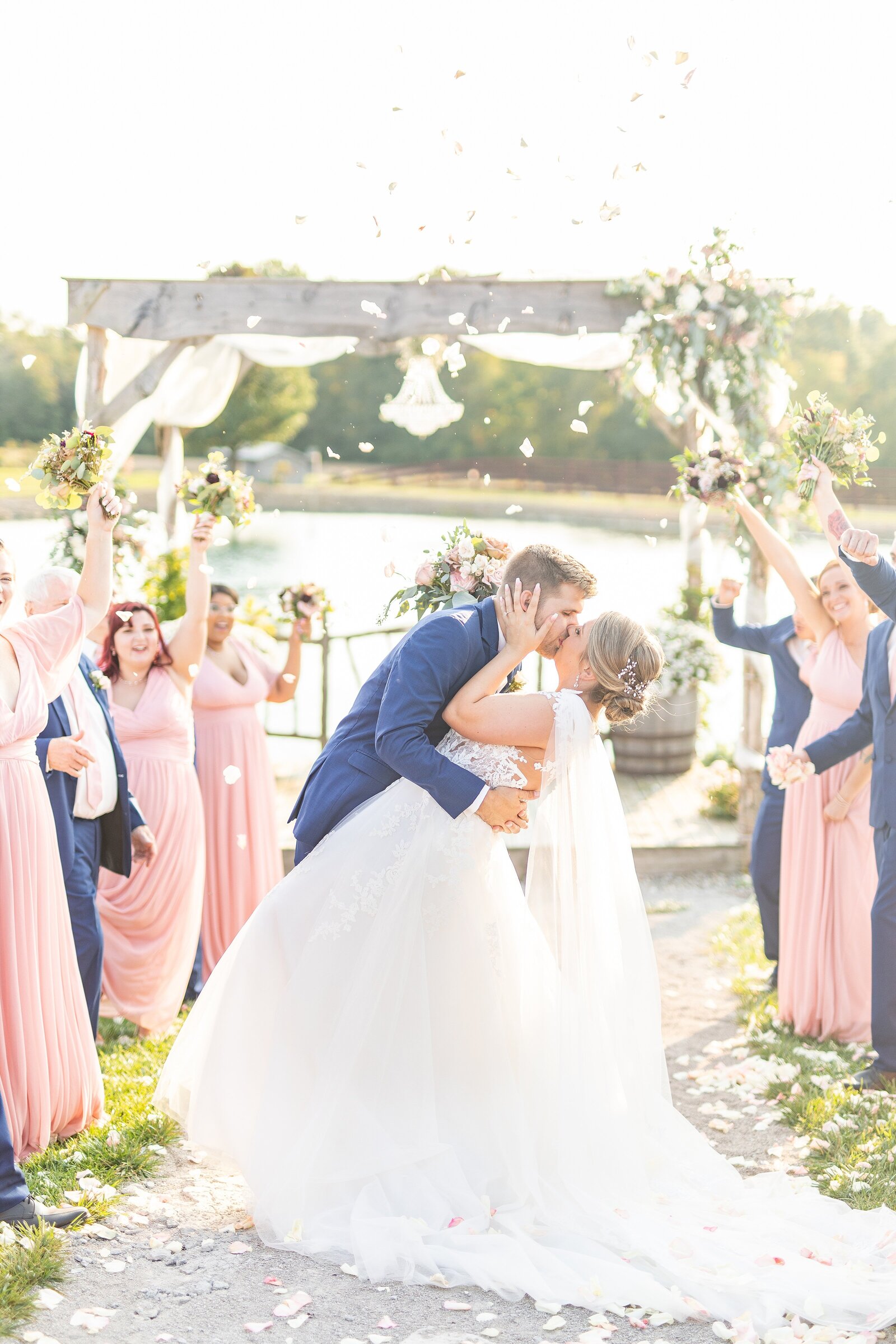 Bride wearing lace A-line gown kissing groom wearing navy suit in front of ceremony arbor with wedding party cheering and throwing white rose petals.
