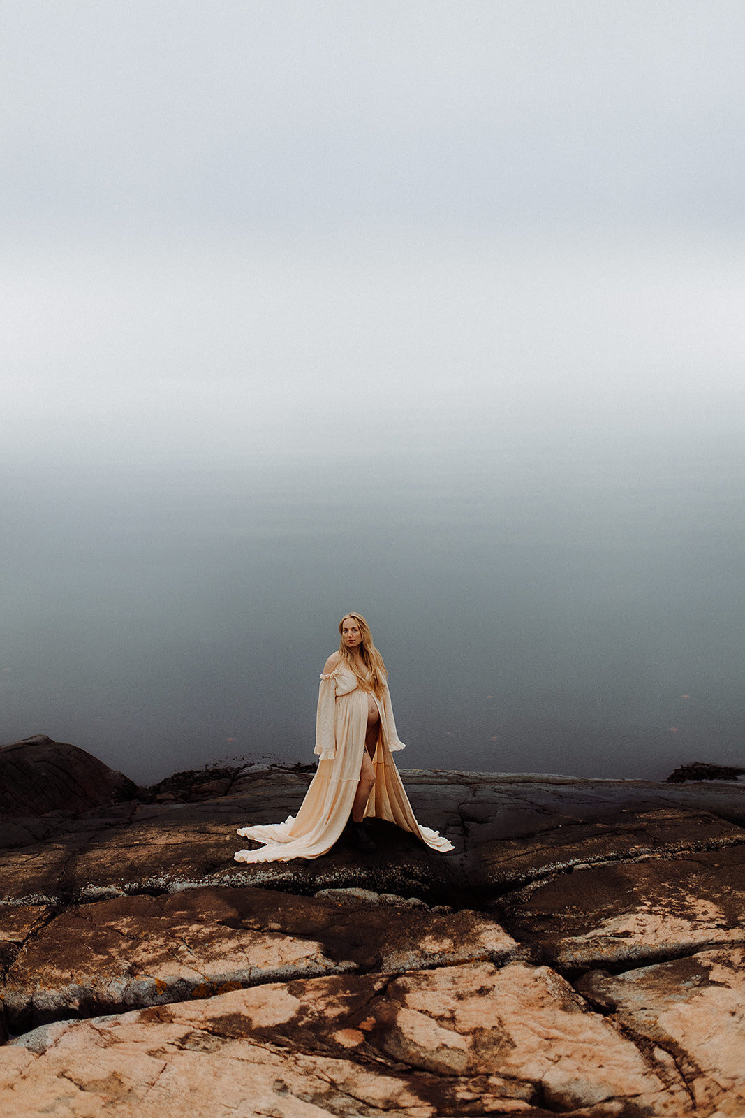 en gravid mamma står vid havet i en öppen gravidklänning som visar hennes mage