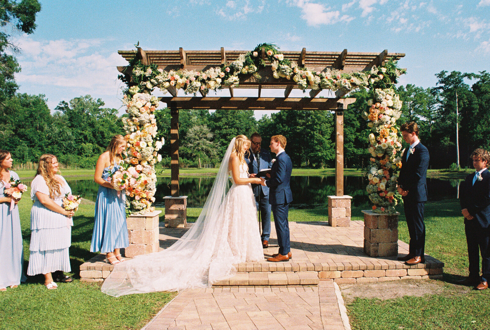 Bride and groom hold hands at the alter before saying their "I do's" to each other under an outdoor pergola with flowers