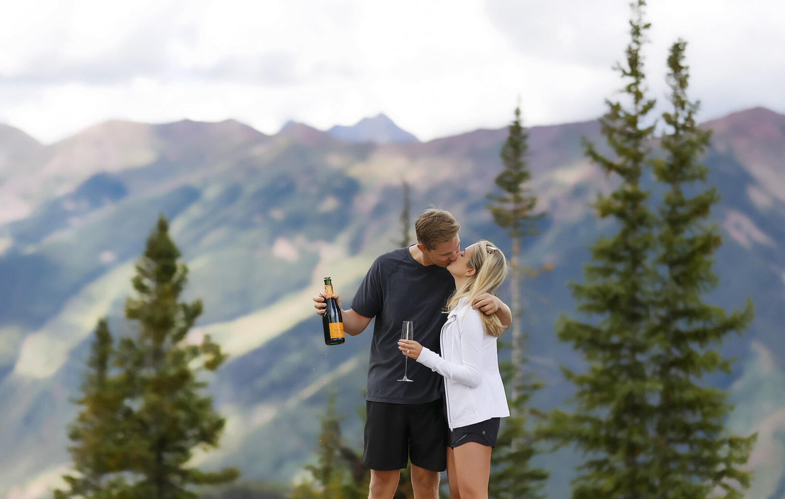 A young man and woman share a kiss and open a bottle of champagne after his surprise proposal  on Aspen Mountain in Aspen, Colorado.