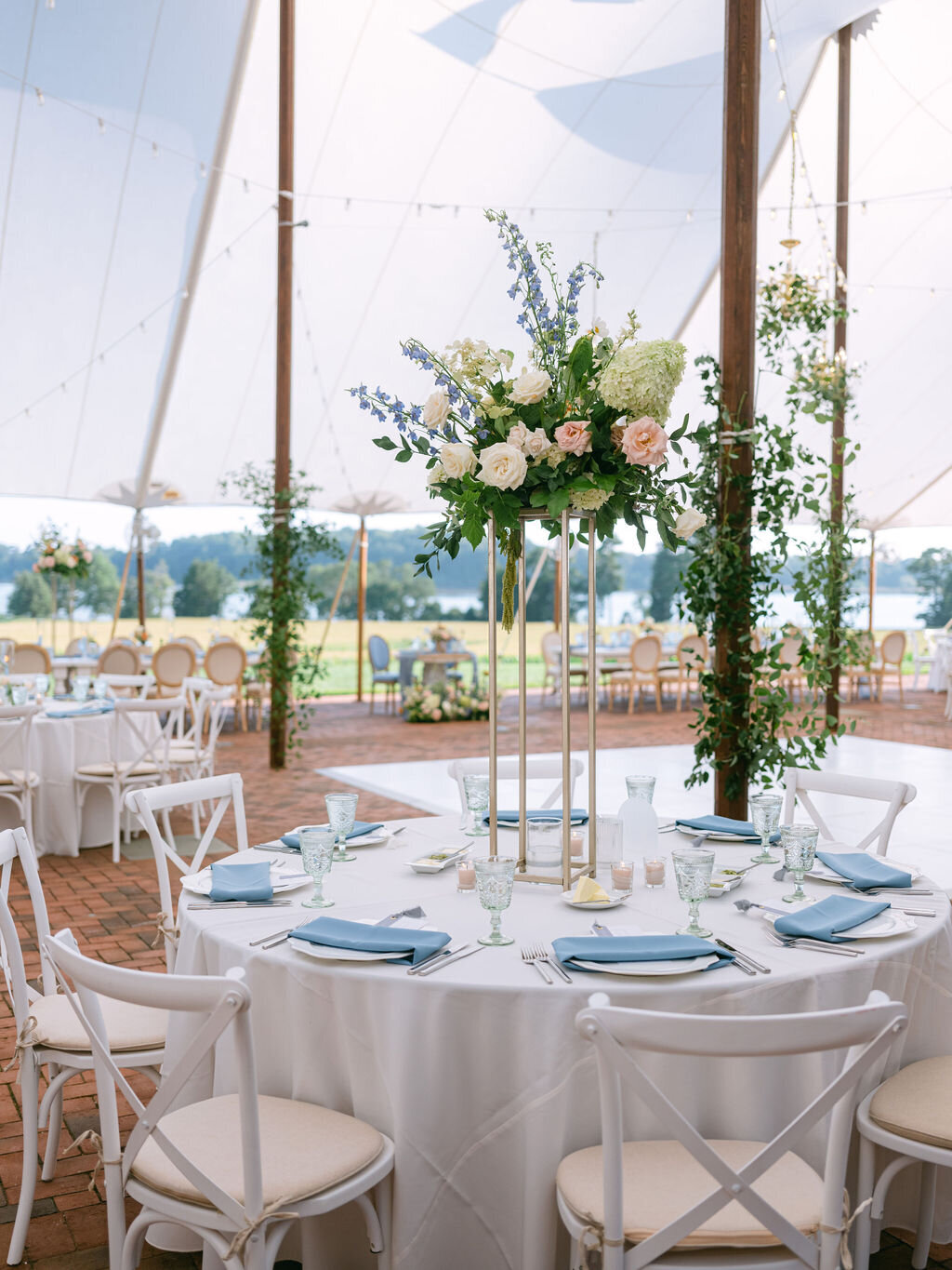 Tented reception at Brittland Estate with smilax greenery on tent polls and arrangement on a gold stand at a round guest table with florals including green hydrangea, blue delphinium, toffee roses, blush garden roses, and green hanging amaranthus.