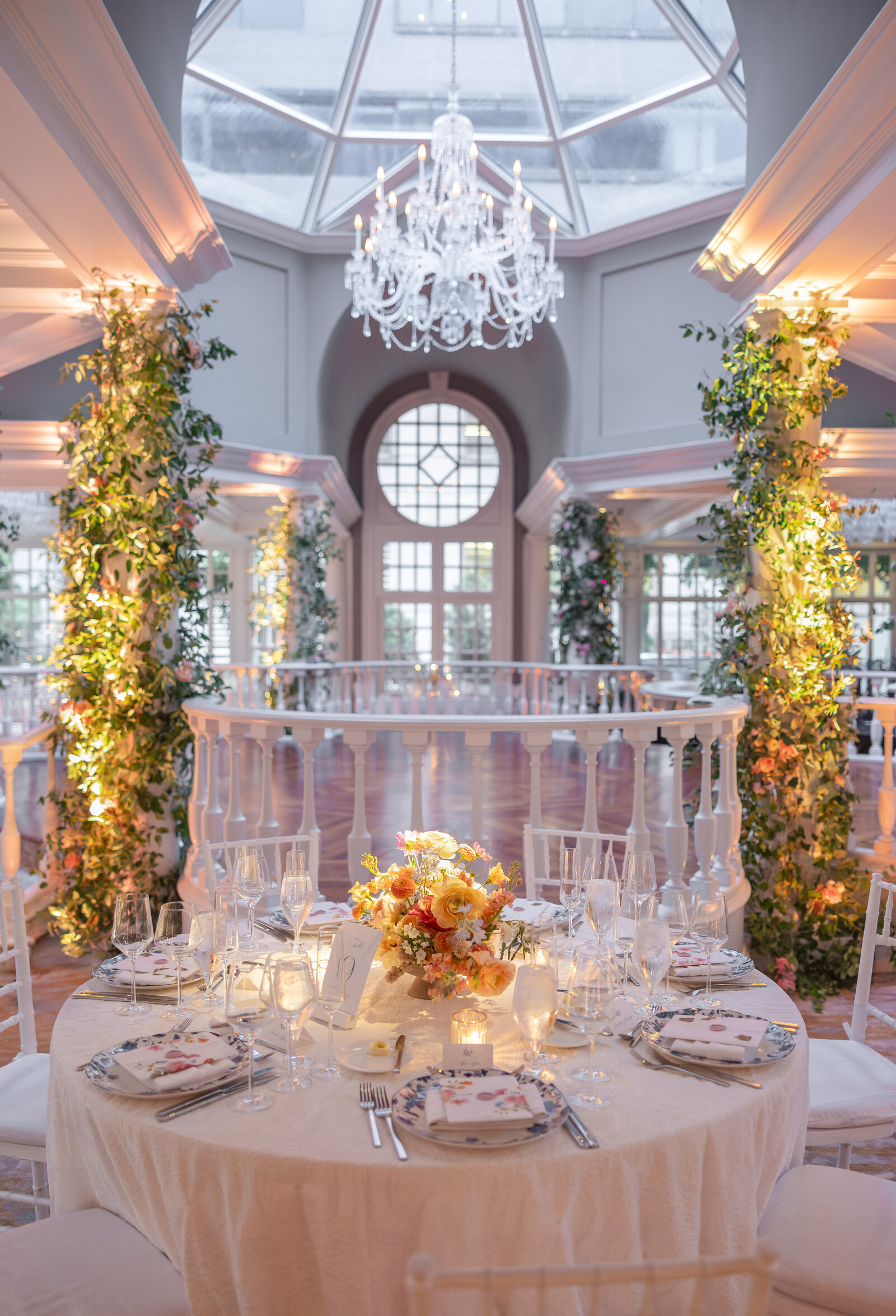 A beautifully set dining table with floral centerpieces and elegant tableware, situated under a grand chandelier in an ornate, light-filled room. The space is adorned with vertical greenery and large windows, creating a sophisticated ambiance.