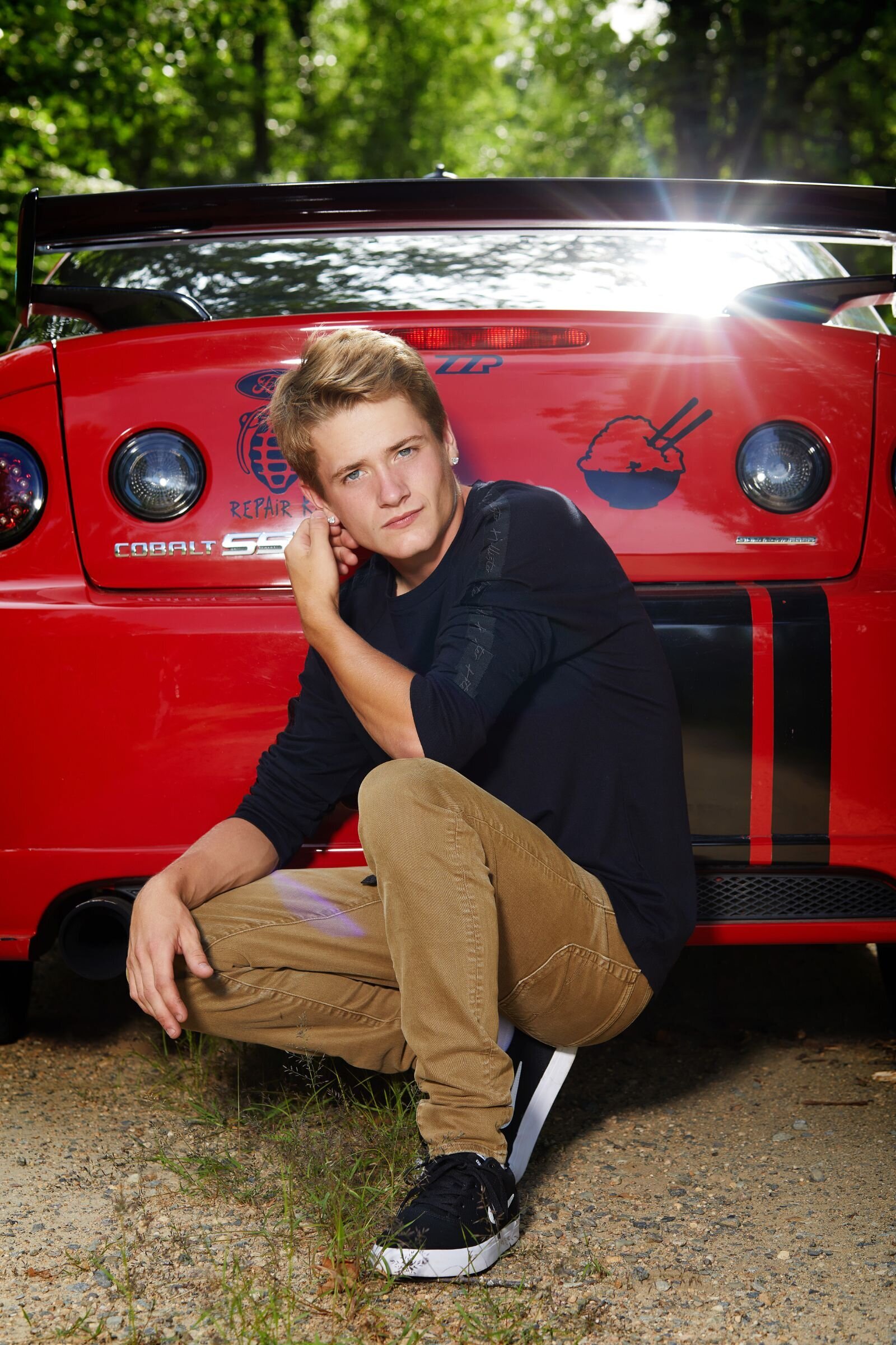 HIgh School senior guy crouching down in front of his red car on a gravel road with a serious expression.