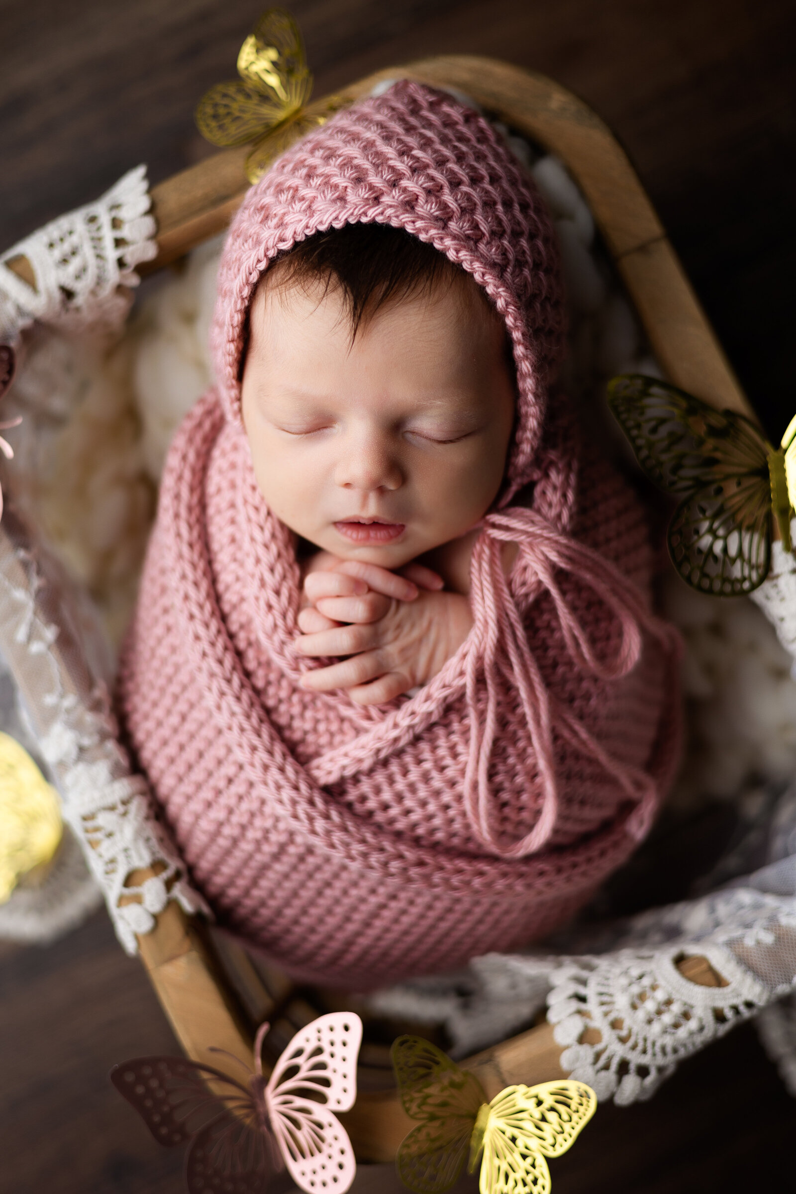 Newborn baby girl swaddled in a pink knit wrap with matching bonnet and posed in a wooden crate surrounded by butterflies