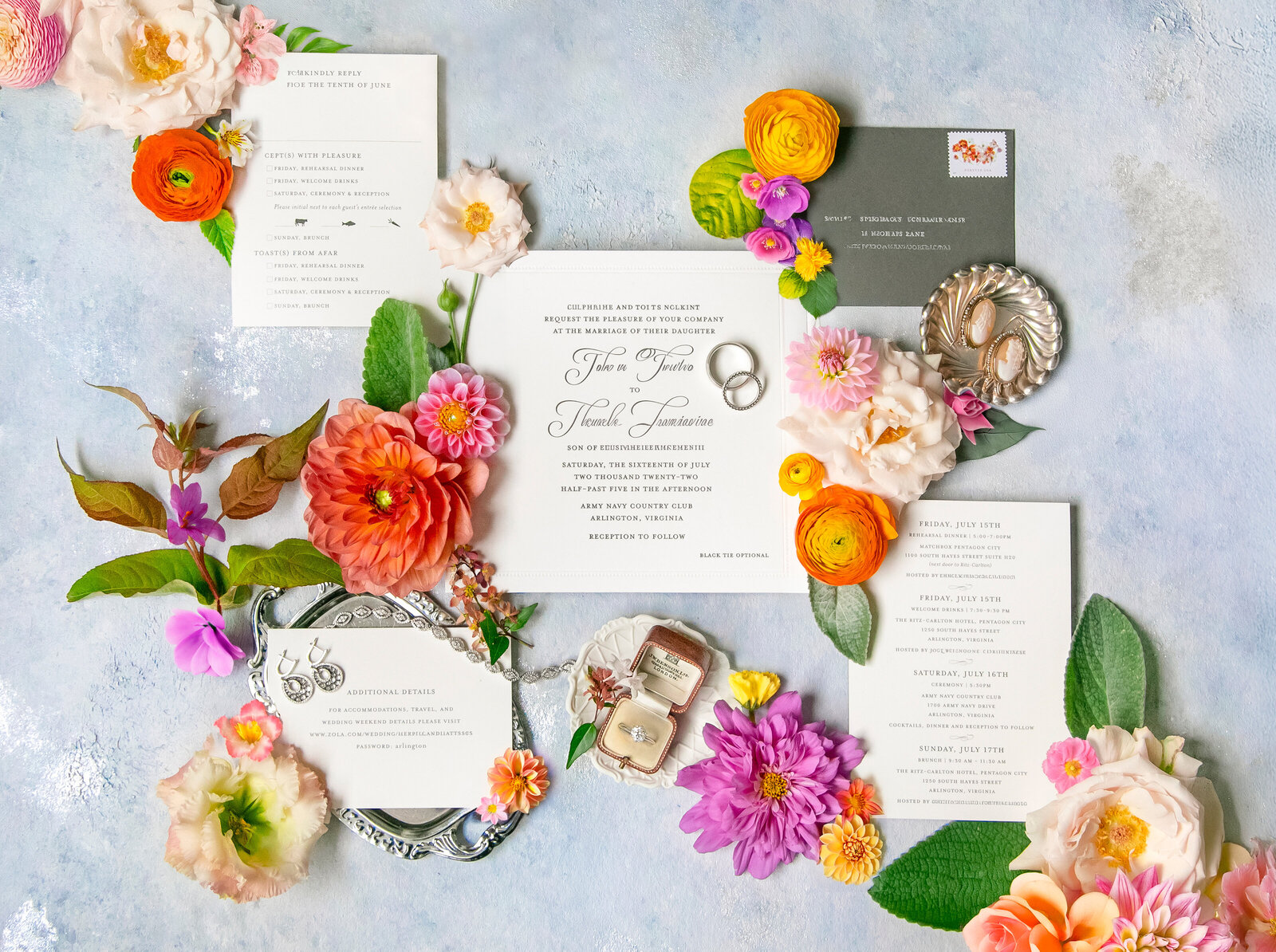 A wedding invitation set surrounded by vibrant flowers, greenery, and decor elements like vintage keys and rings on a light blue surface. The arrangement includes various stationery pieces with elegant typography.
