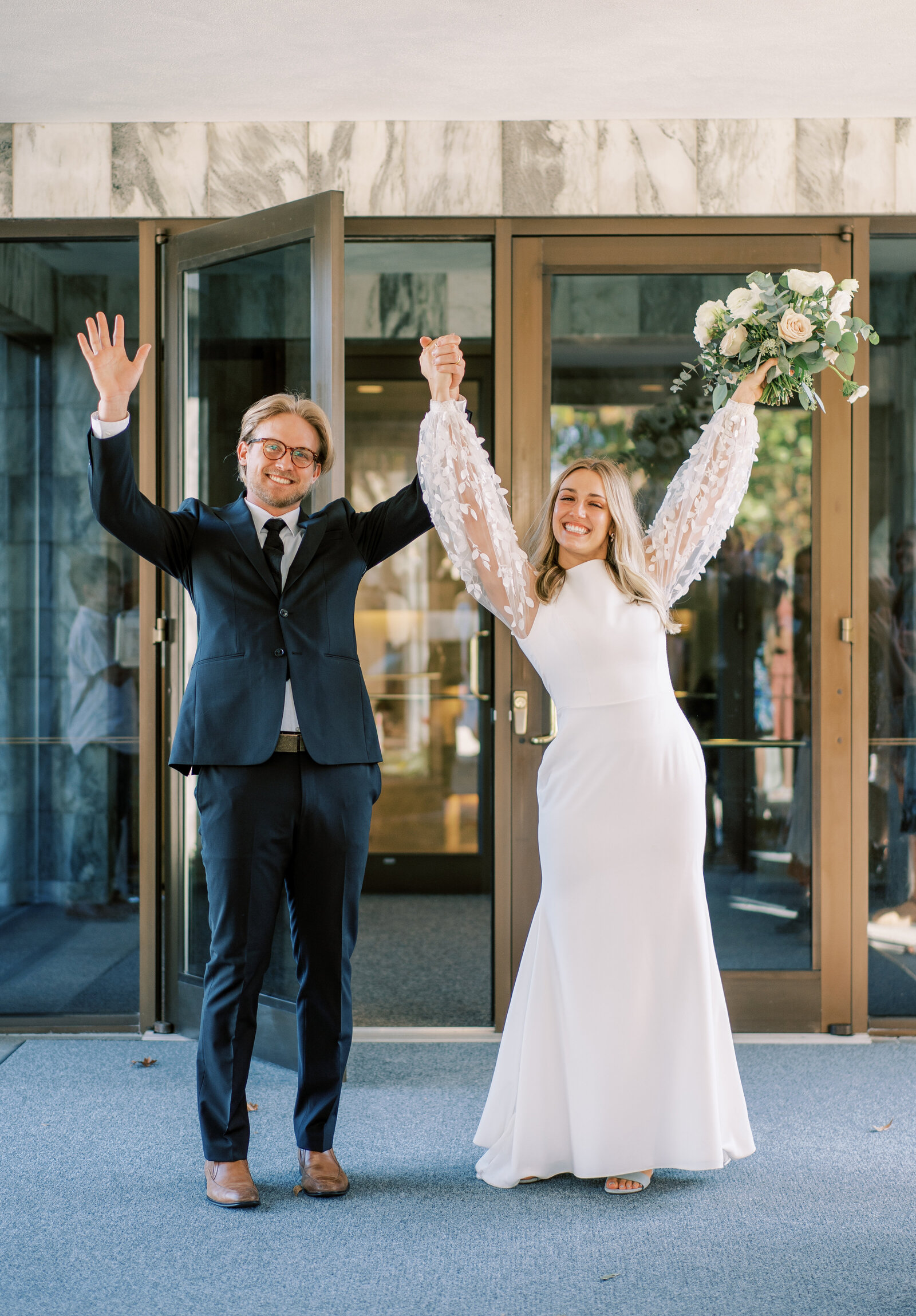 Portrait of bride and groom in a white wedding gown and black suit standing with glass doors outback.