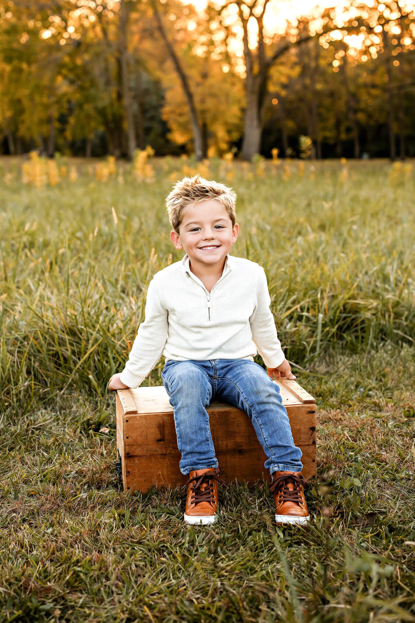 A smiling child with short blond hair sits on a wooden crate in a grassy field, perfectly capturing the essence of outdoor portraits. Dressed in a white long-sleeve shirt, blue jeans, and brown sneakers, they are framed by tall grass and trees with autumn foliage under a sunset sky.