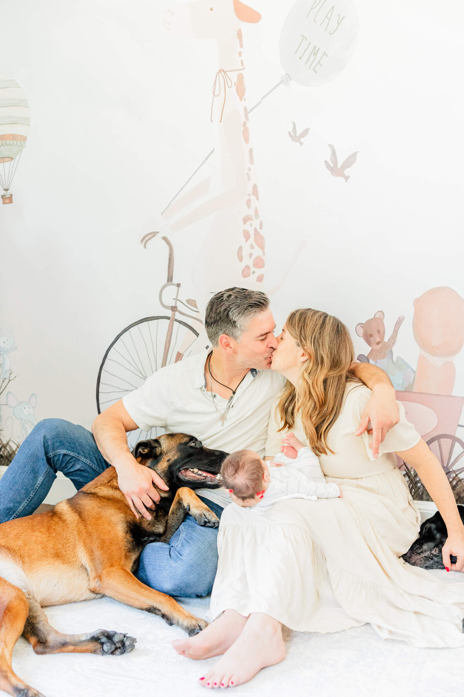 Mom and dad share a kiss while sitting on the floor in front of an animal circus nursery wall with two dogs laying next to them