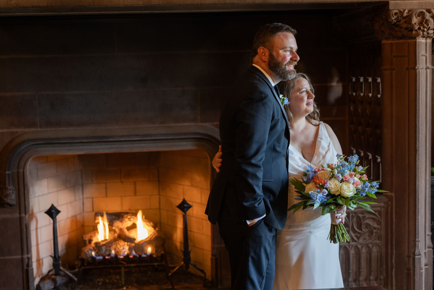 A bride and groom are standing in front of a fireplace.
