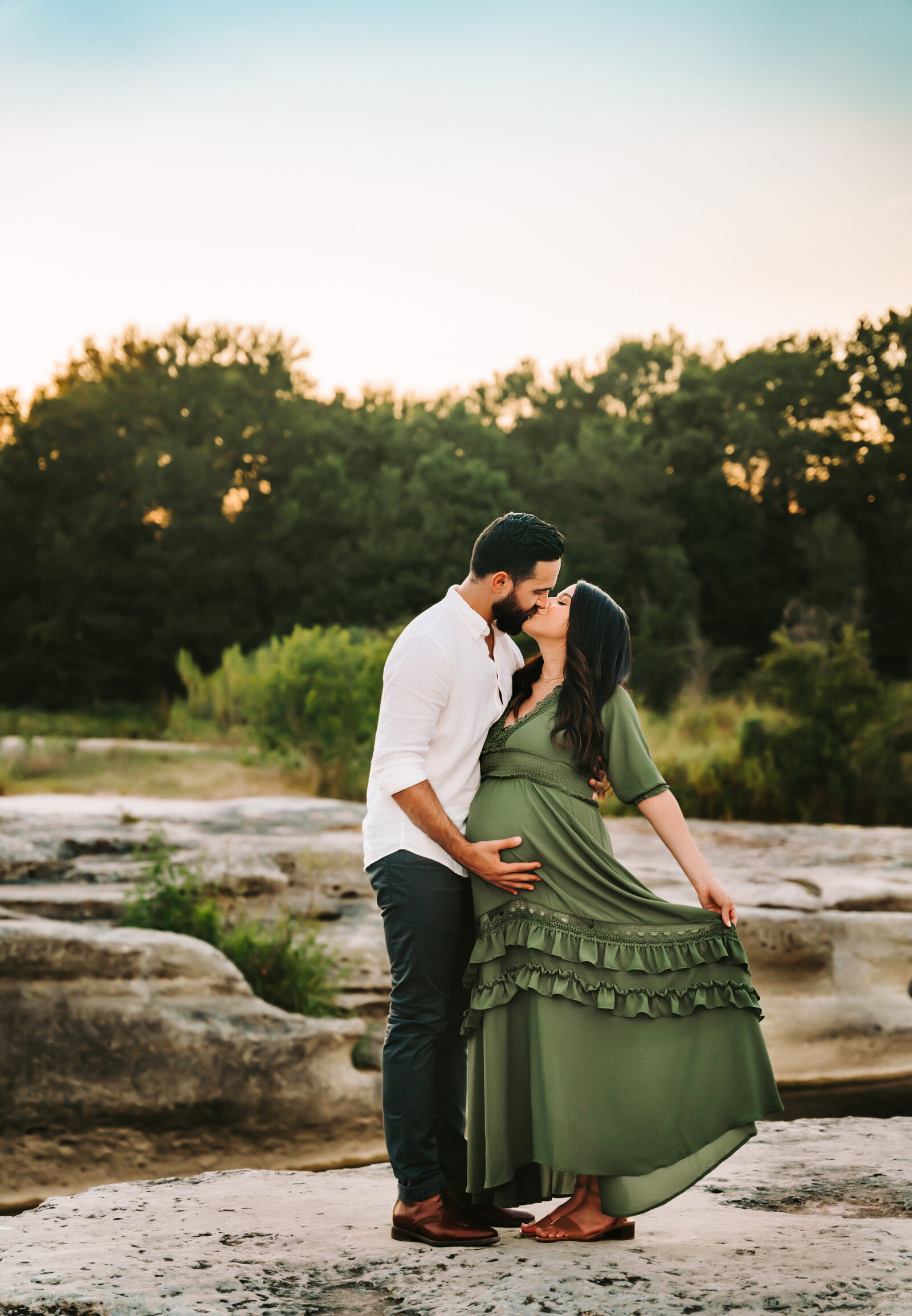 Maternity Photographer, a man and his pregnant wife kiss, she holds her dress out near the river