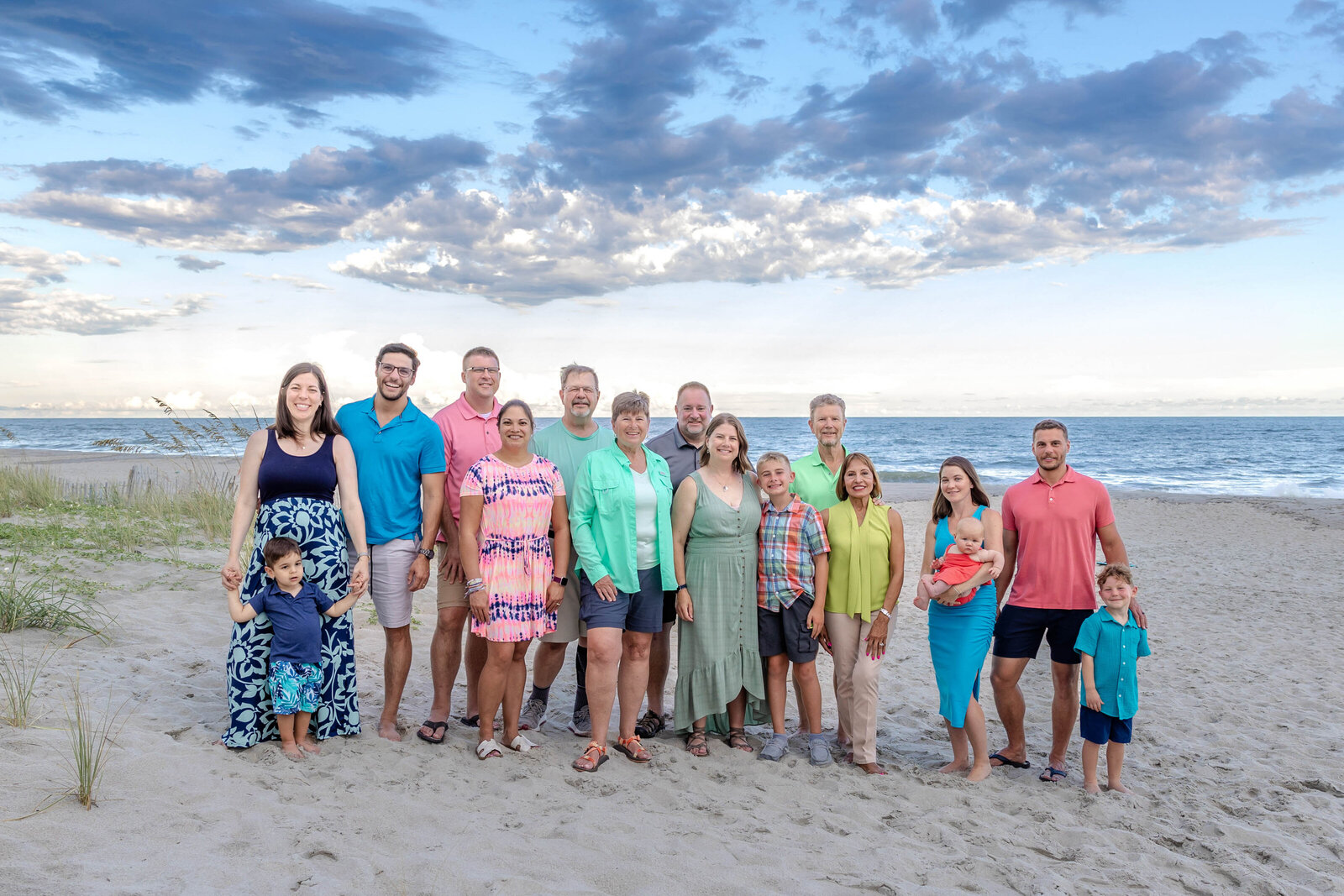 A large family stands together on the beach, dressed in vibrant, colorful outfits. The bright sky and ocean add to the cheerful atmosphere, ideal for destination family photographer NC, capturing lively and joyful family connections.