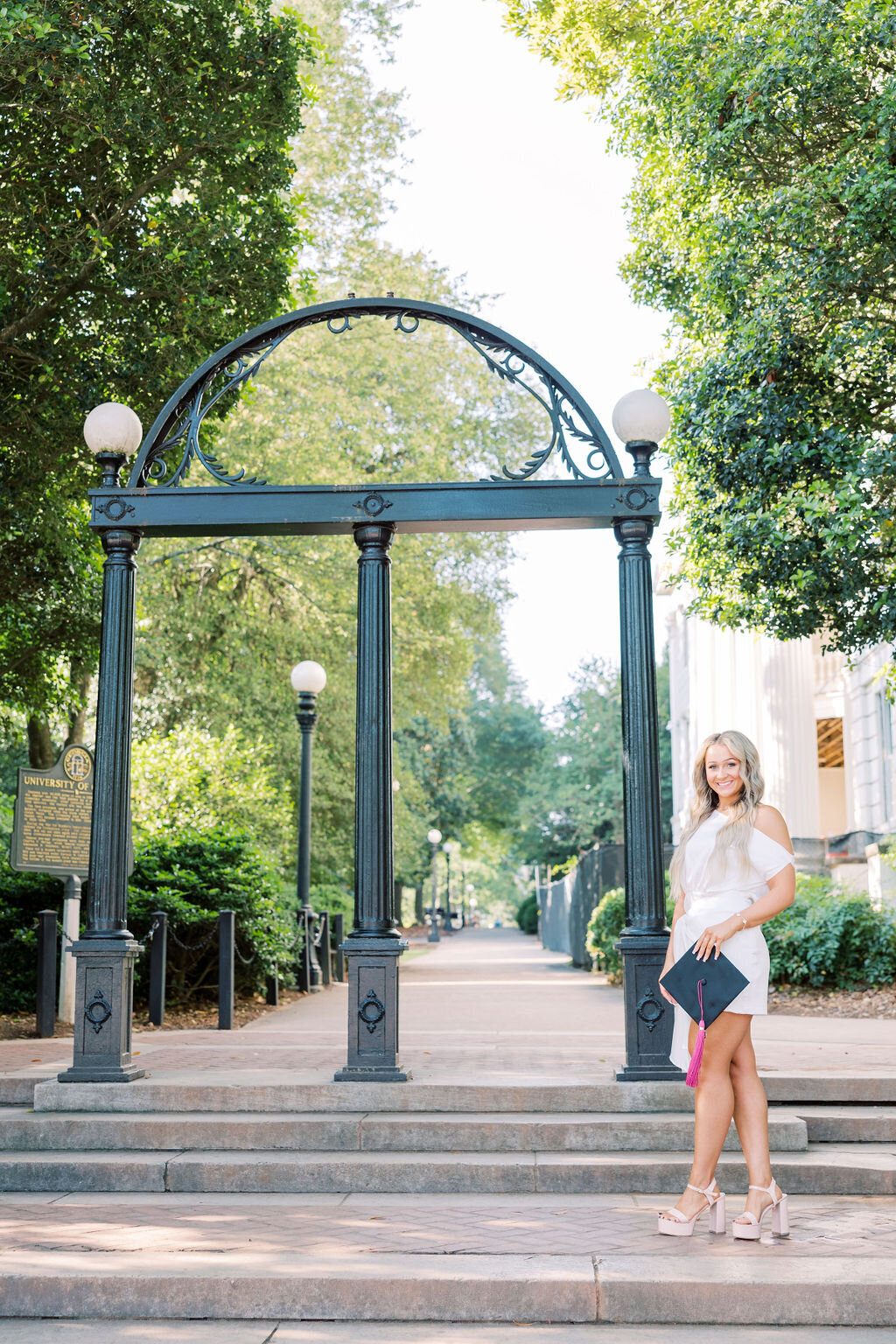 Girl holds graduation cap standing under UGA Archway