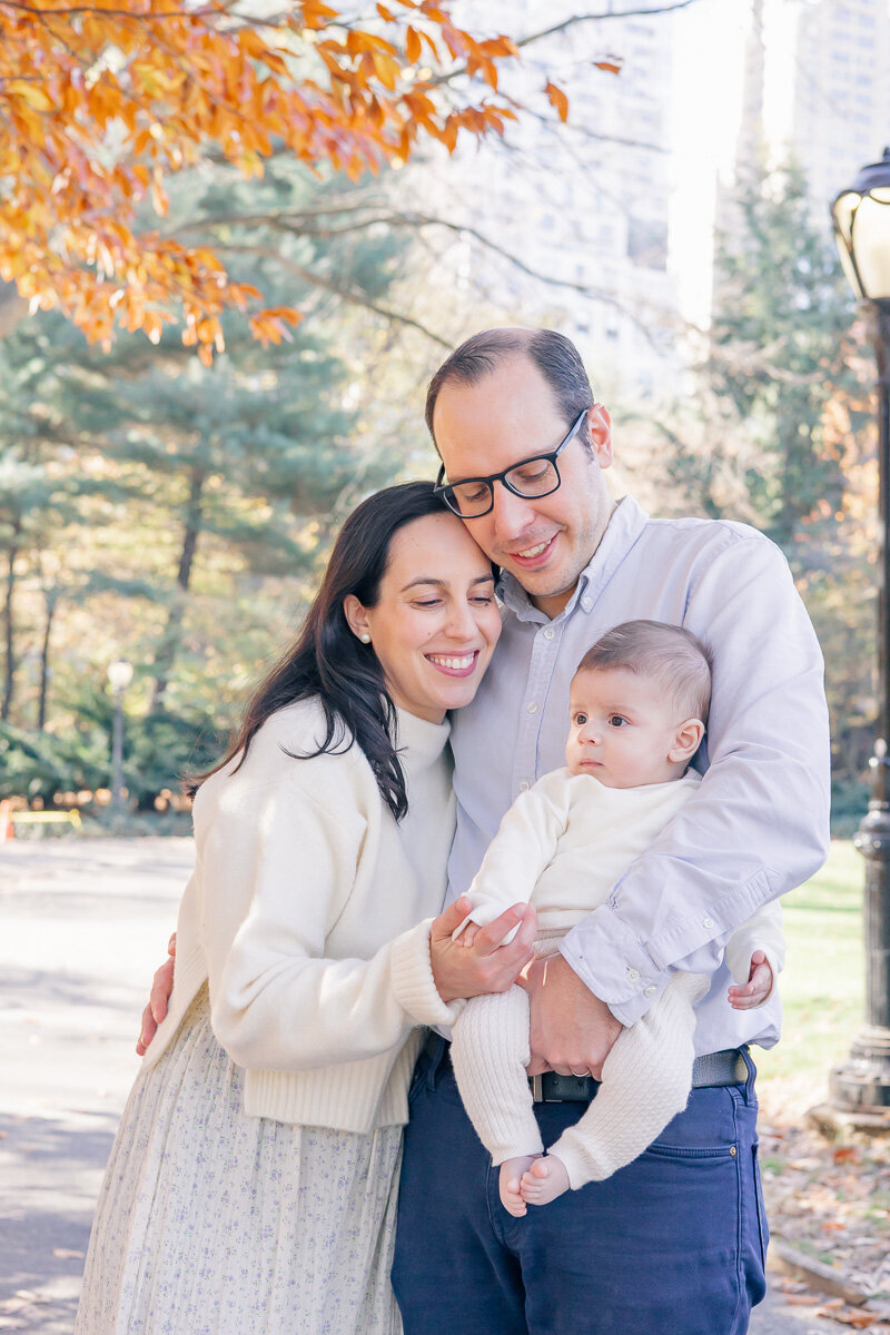 Candid family photo in a Park featuring a mom, dad, and baby snuggled close under autumn trees. The parents smile with eyes closed, soaking in a peaceful moment together while their baby looks out curiously. A soft, love-filled image captured by a Greenwich family photographer who documents heartfelt, slow-living moments for families who cherish connection.