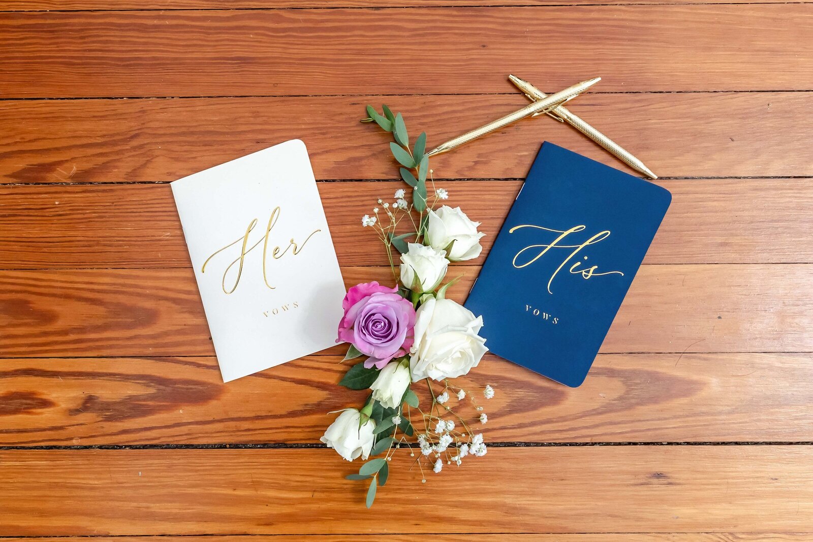 His and Her vow books with gold lettering are displayed with white and purple roses and golden pens, captured on natural wood flooring during a Coatesville, PA wedding, showcasing personalized wedding details.