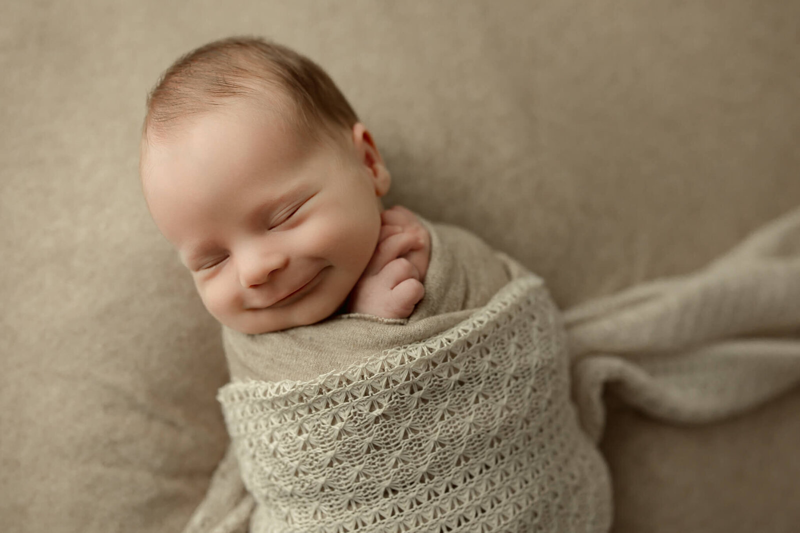 Baby boy with a smile swaddled in a monochromatic tan set up during his newborn photos