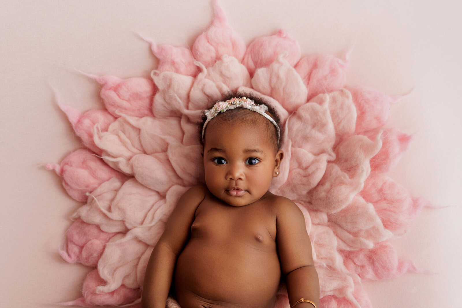 A beautiful baby with dark skin and bright eyes lies on a soft pink blanket shaped like petals, resembling a flower. The baby wears a delicate headband adorned with flowers and gazes curiously at the camera, creating a serene and enchanting scene.