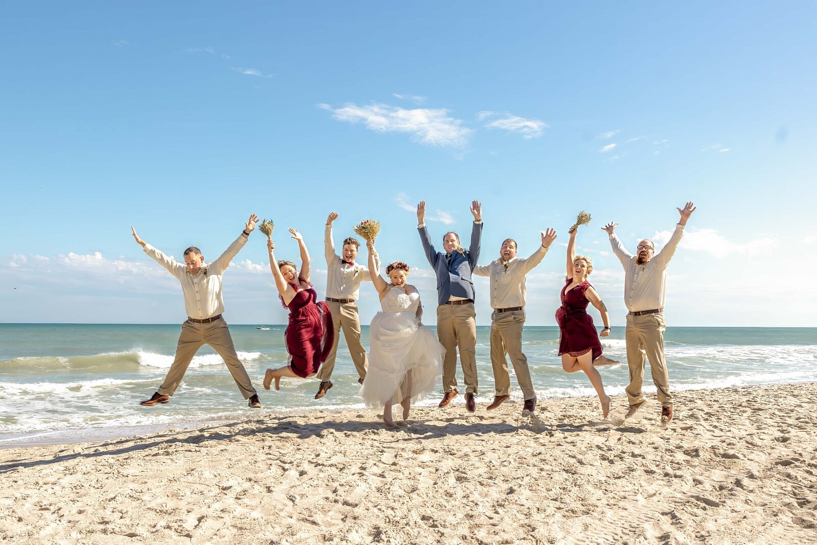The bride, groom, and wedding party jump with excitement on the sandy shores of Carolina Beach, NC, under a bright blue sky, blending coastal charm with fun and candid moments.