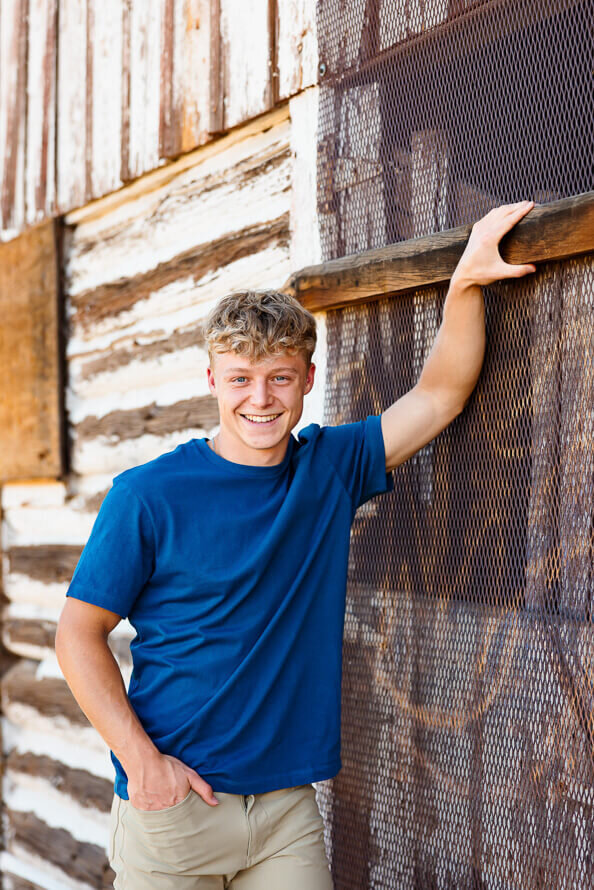 Senior boy in blue holds on to the barn at Hildebrand Ranch Park in Colorado.