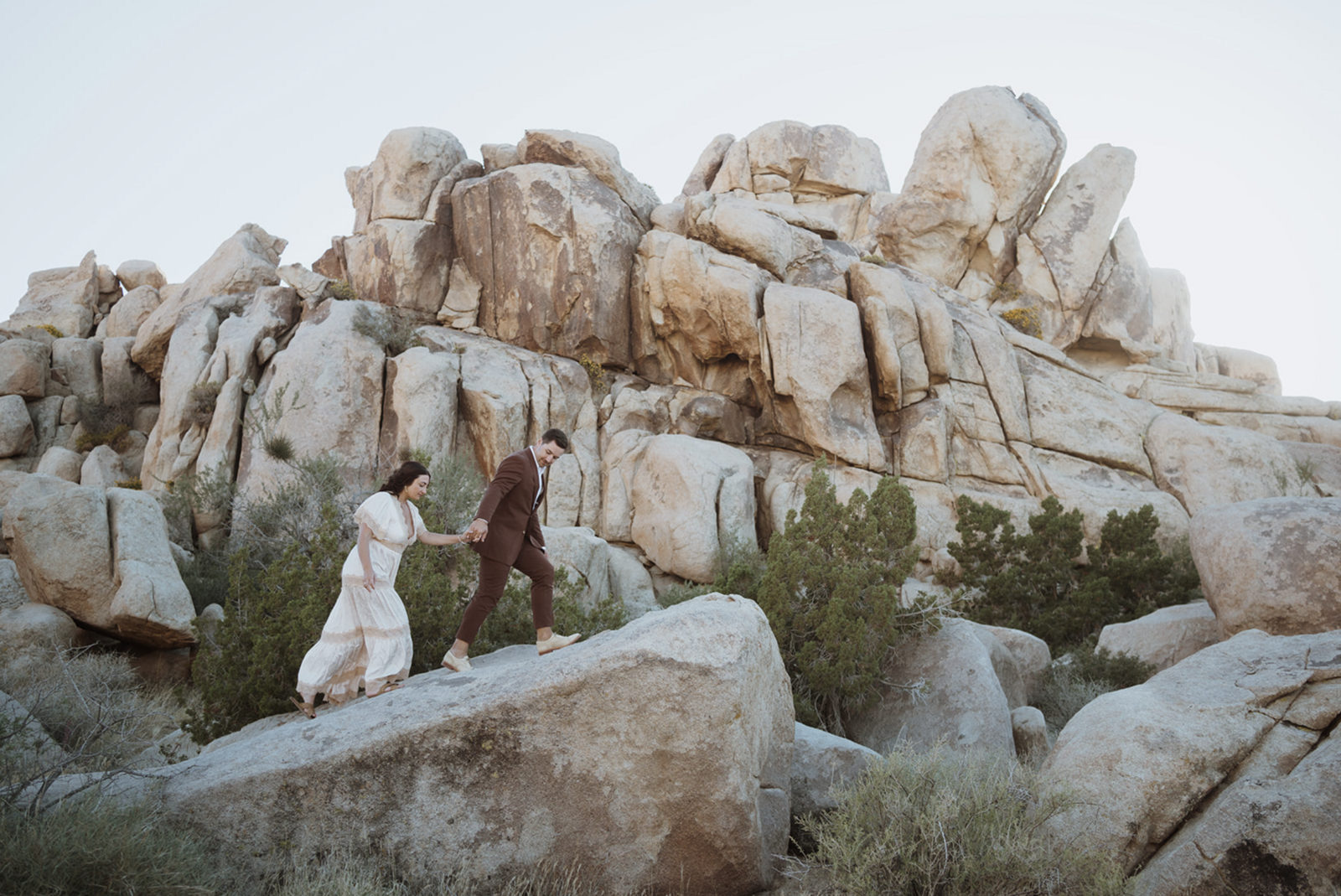 the bride is getting lead by the groom up a rock. they are hiking in their wedding attire - the groom is in a maroon suit and the bride is in a white and tan dress. they are surrounded by rocks and bushes. the sun is setting behind them so it's shaded.