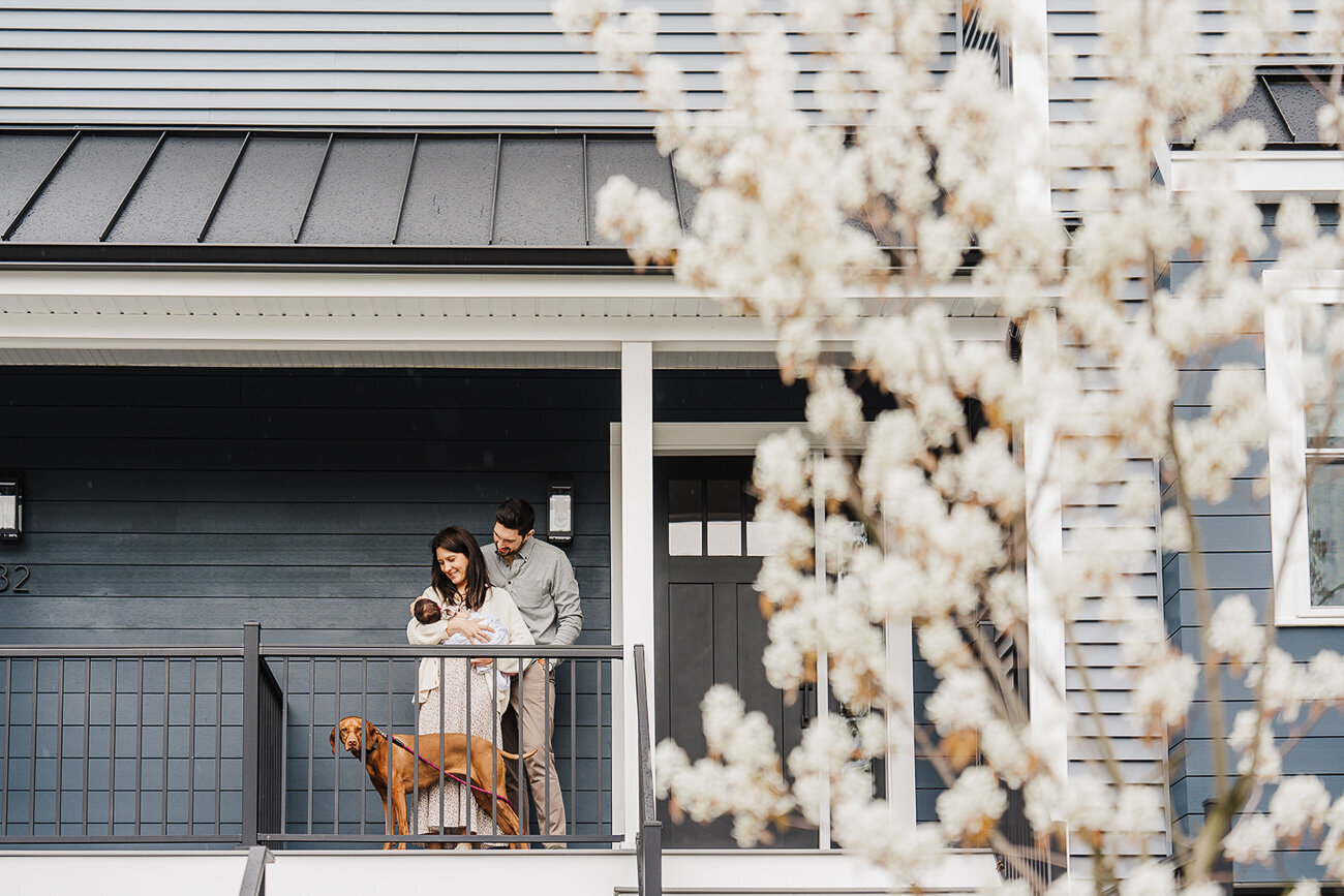 parents hold baby girl and stand on front porch with blossoming tree