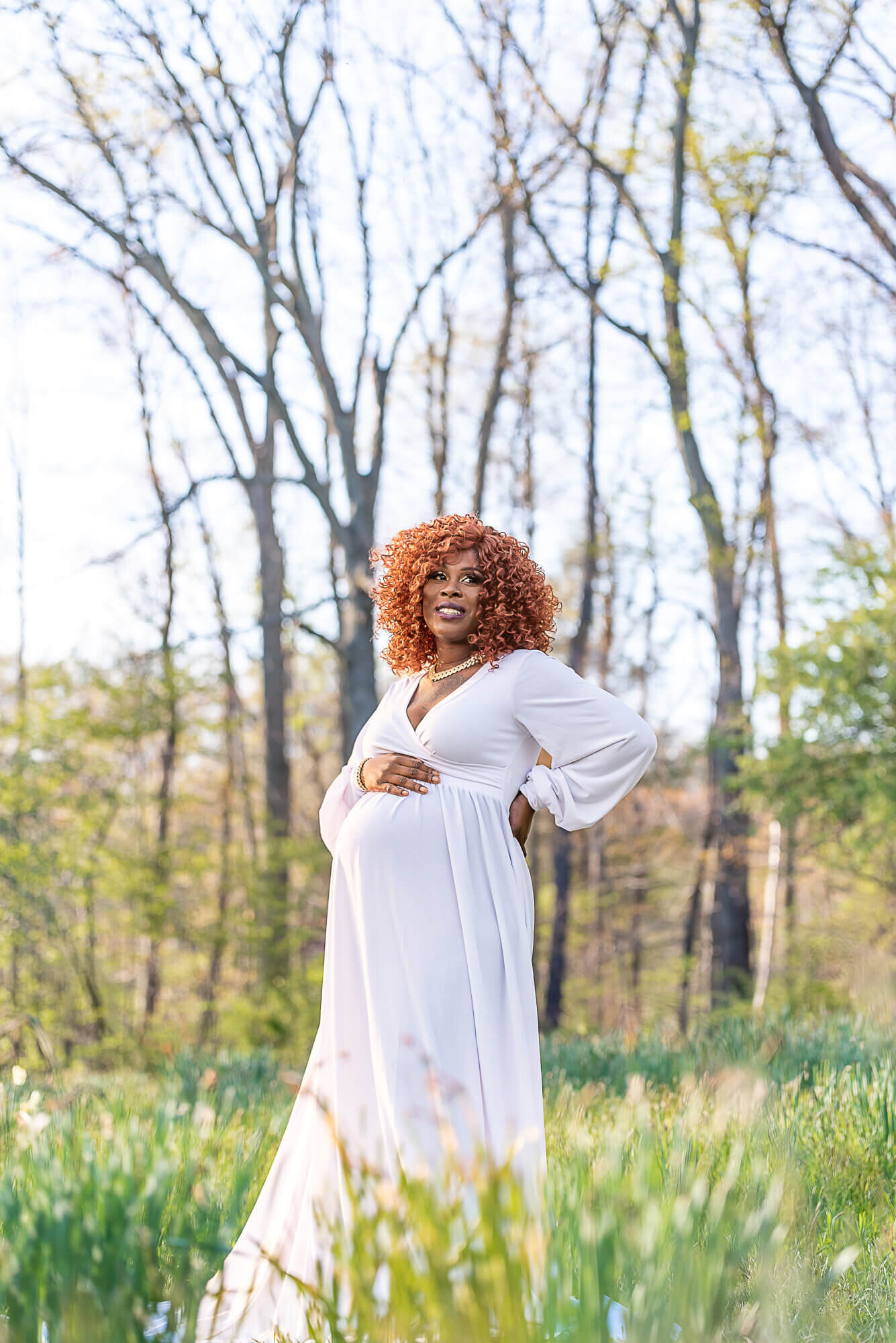 A pregnant woman in a flowing white dress stands in a lush, sunlit field. She smiles while holding her baby bump, surrounded by tall trees in the background.