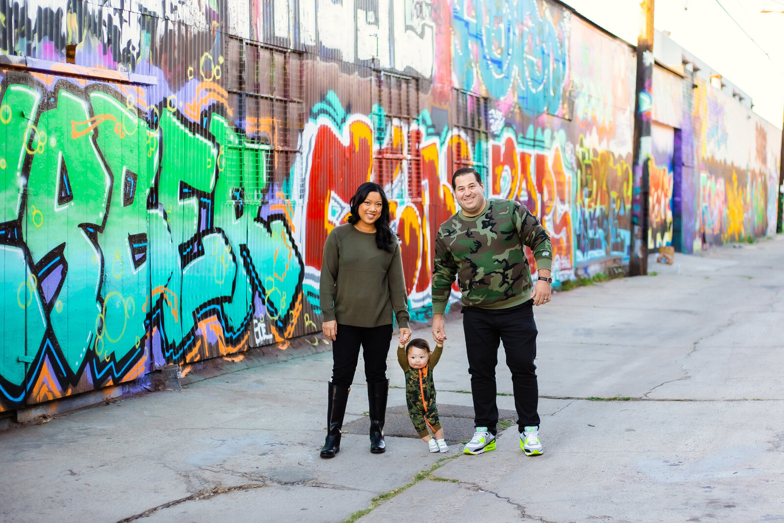 Family Photographer, a young mother and father walk with their baby near a graffiti colored wall