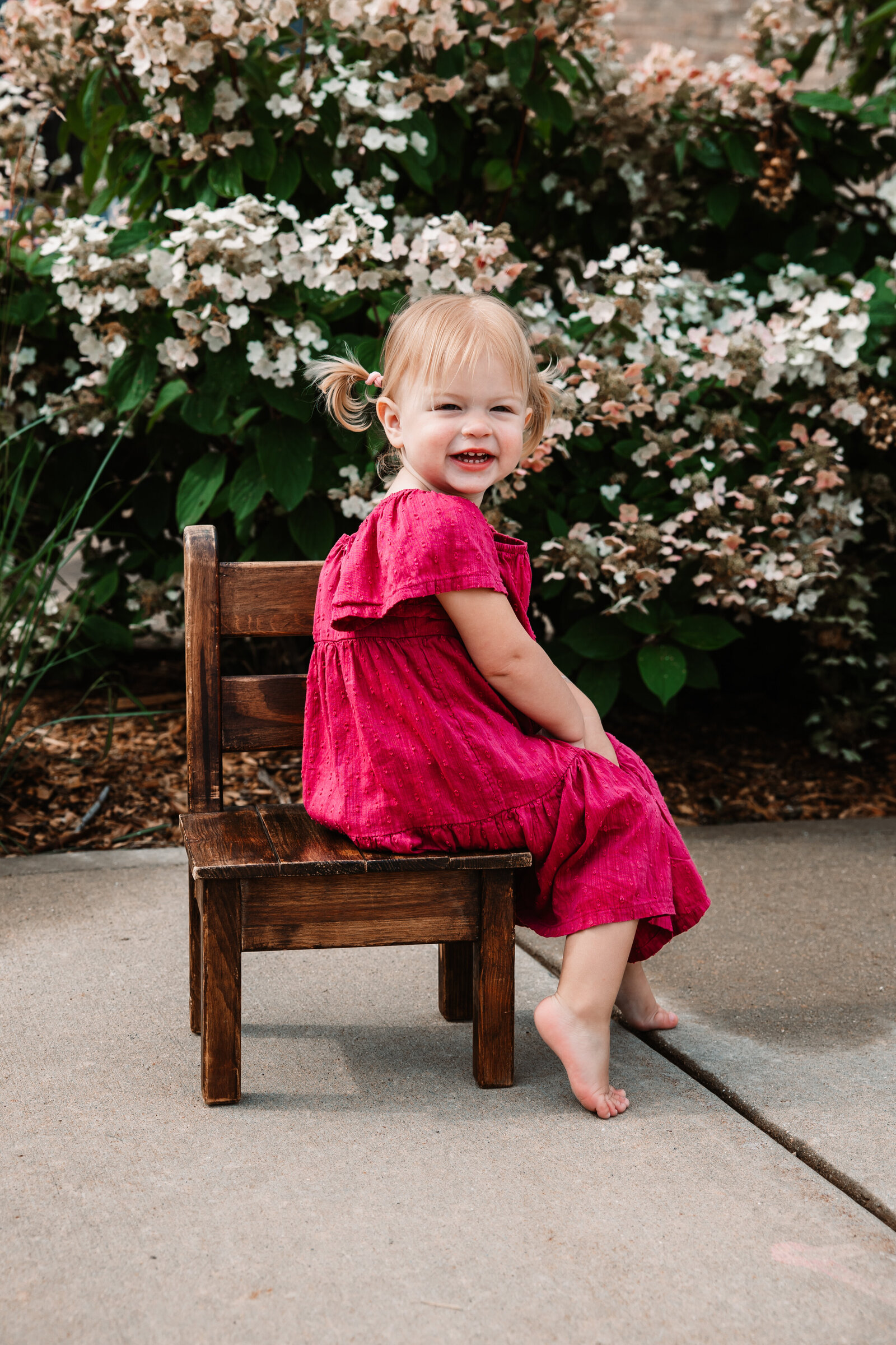 A toddler girl sits sideways on a wooden chair in a flower garden sidewalk