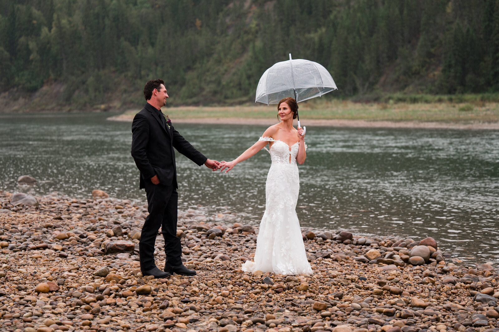 bride and groom walk along the beach by the river in the rain, looking at each other