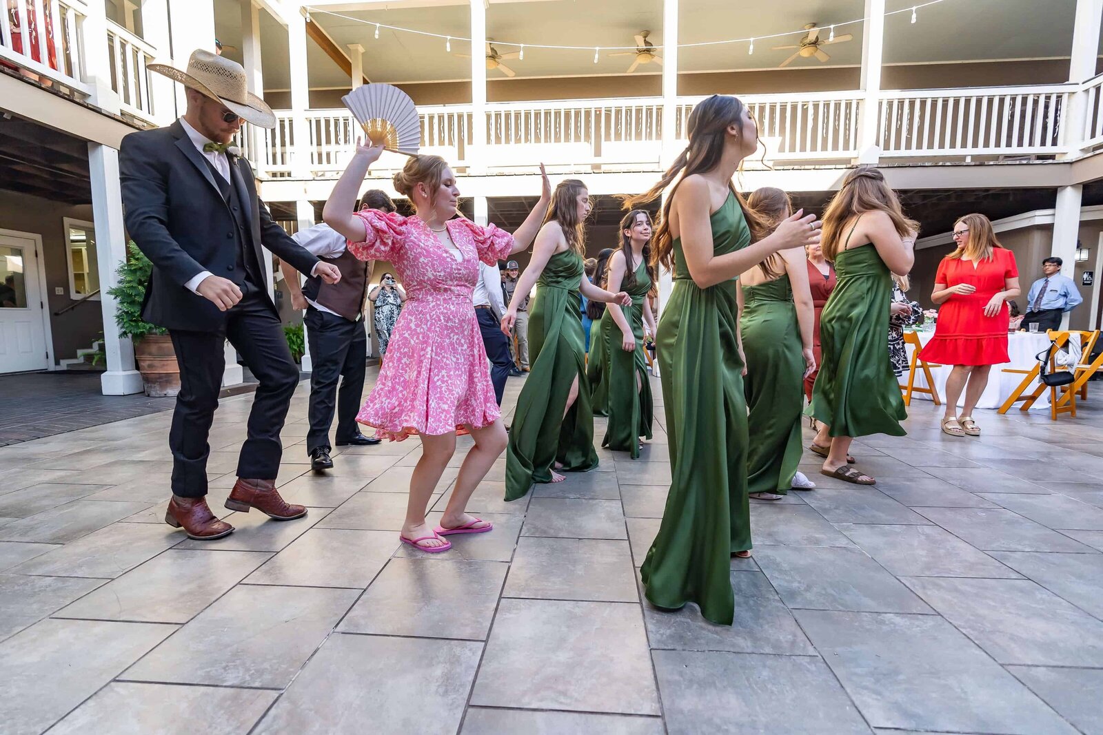 A group of people, including women in green dresses and a woman in a pink dress holding a fan, dance together on an outdoor tiled patio at a social event. Others watch from the background.