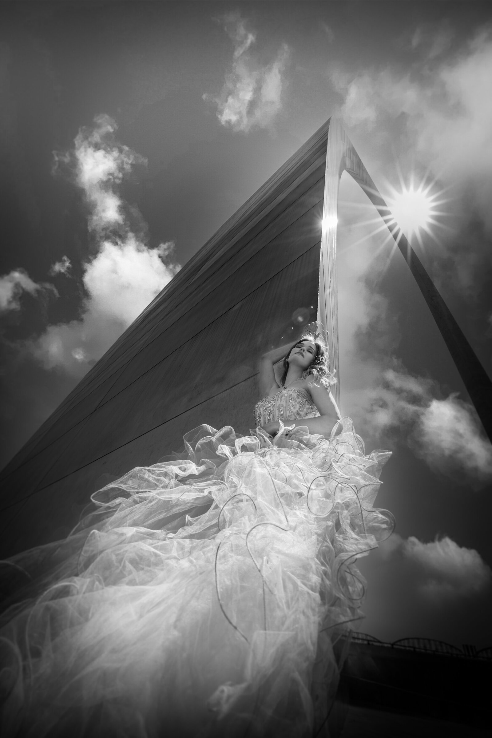 black and white photo taken from the ground looking up at senior in a big ball gown leaning against the gateway arch with the sun overhead