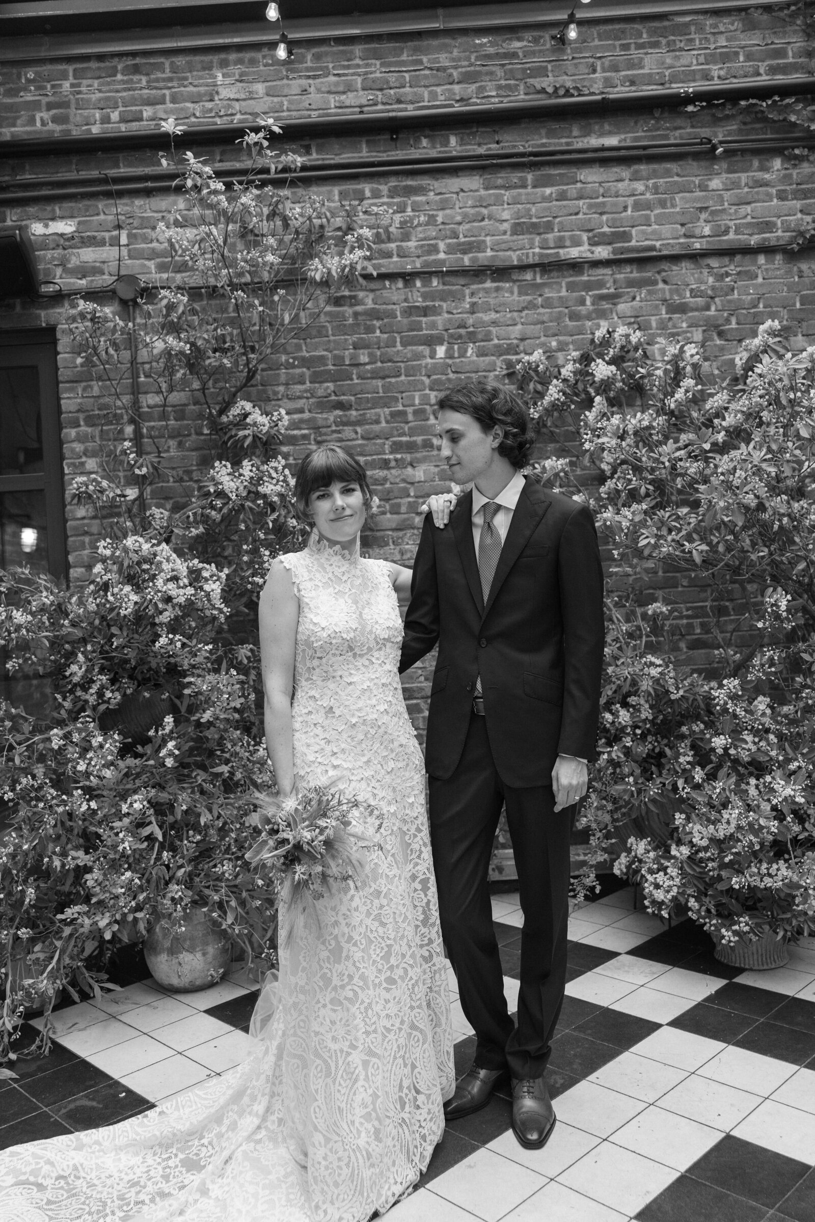 Black & white editorial style wedding portrait of bride and groom casually standing in front of brick wall with florals in NYC 