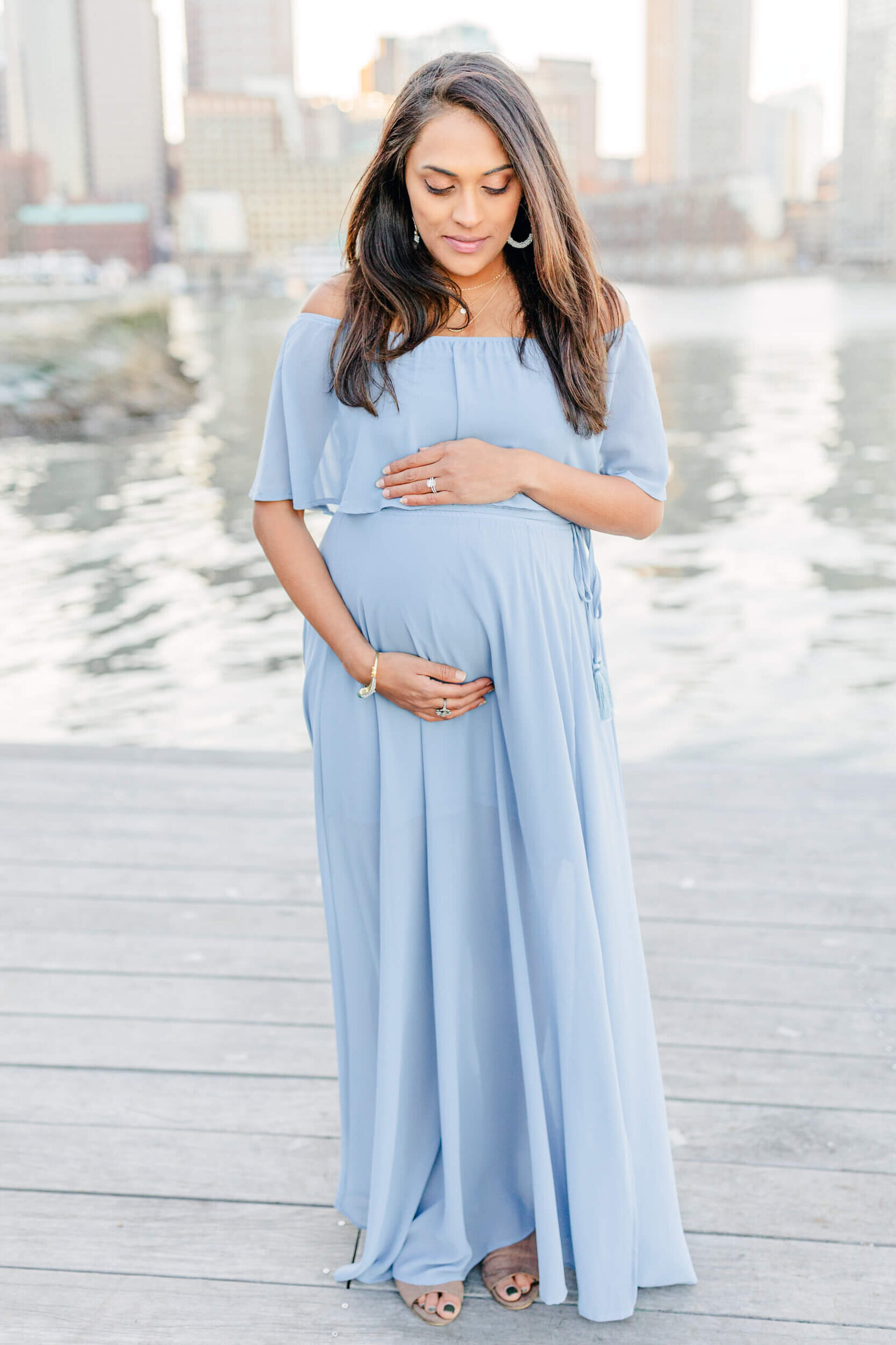 A pregnant woman wearing a pastel blue flowing dress looks down and cradles her bump on a pier at Boston Seaport