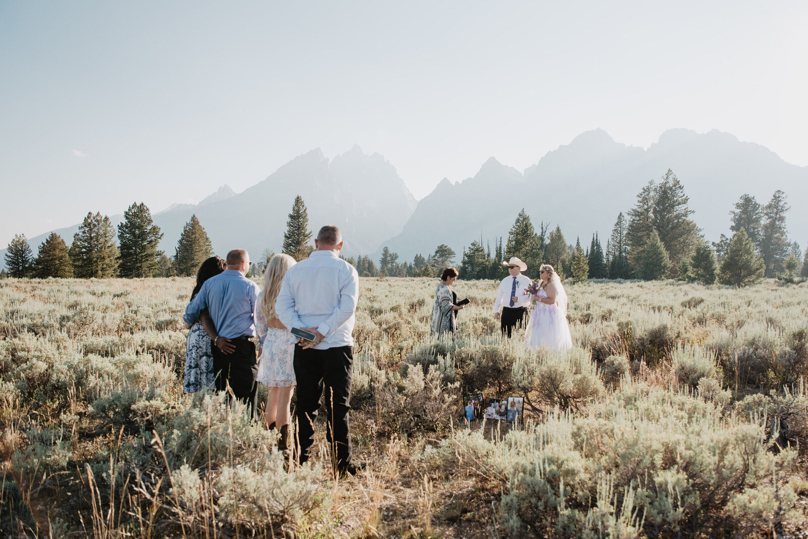 jackson hole wedding photorgaphers captures wedding in a meadow in front of the mountains and a small grove of trees while the sun begins to set as the couples friend watches from a distance for their wyoming elopement