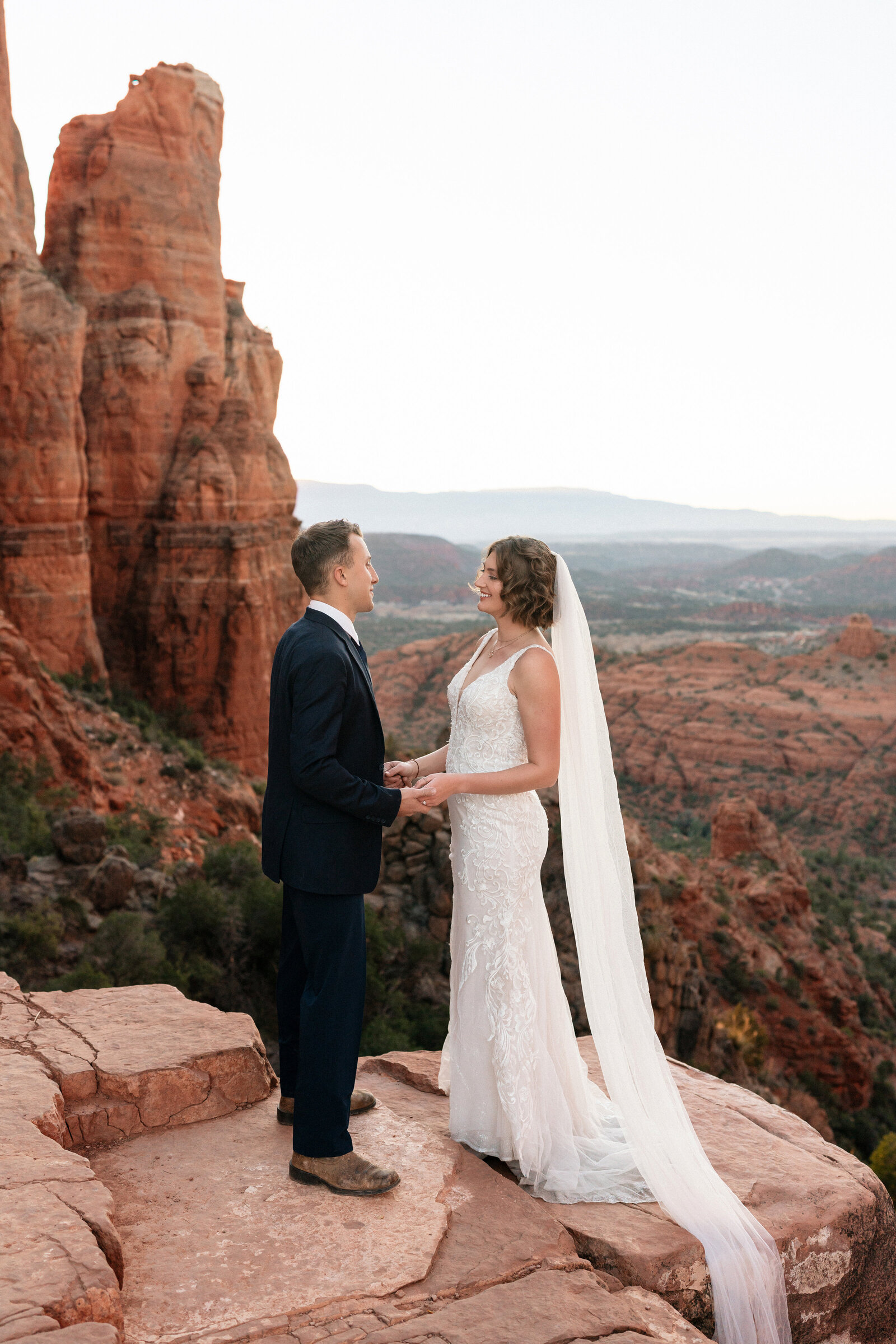 Bride and groom holding hands and exchanging vows on a red rock cliff at Cathedral Rock in Sedona, Arizona, with sweeping desert views and towering sandstone formations in the background.