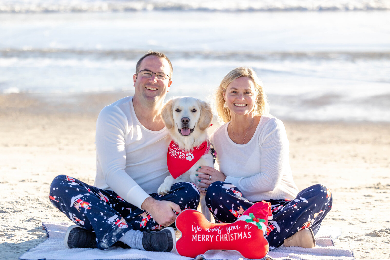 A couple sits on the beach with their dog, dressed in matching festive pajamas. The dog wears a red bandana, and a Christmas pillow adds seasonal charm. Perfect for North Carolina family photography, capturing holiday joy by the ocean.
