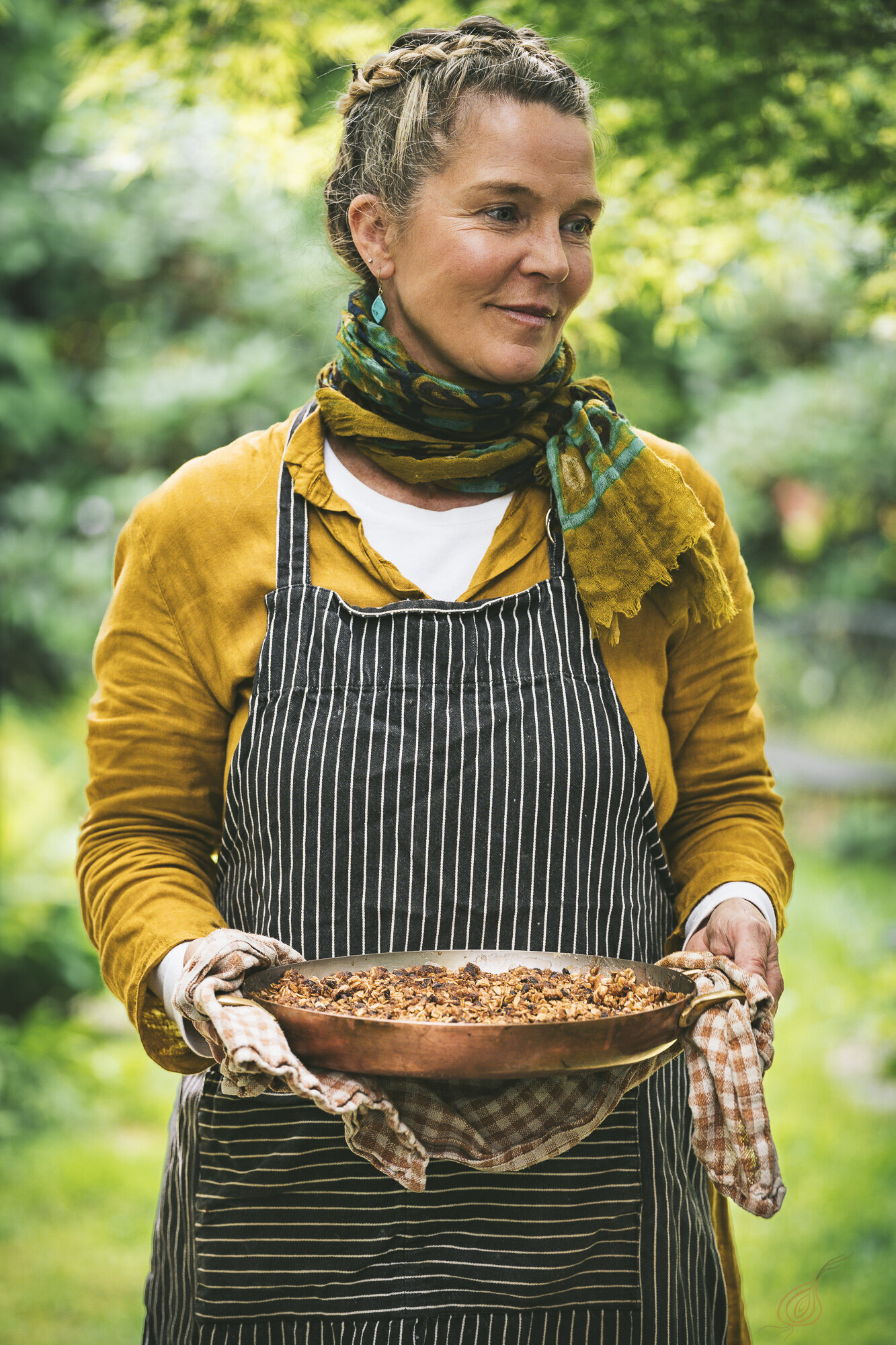 A woman in an apron stands holding a pan filled with delicious food, ready to serve a meal.