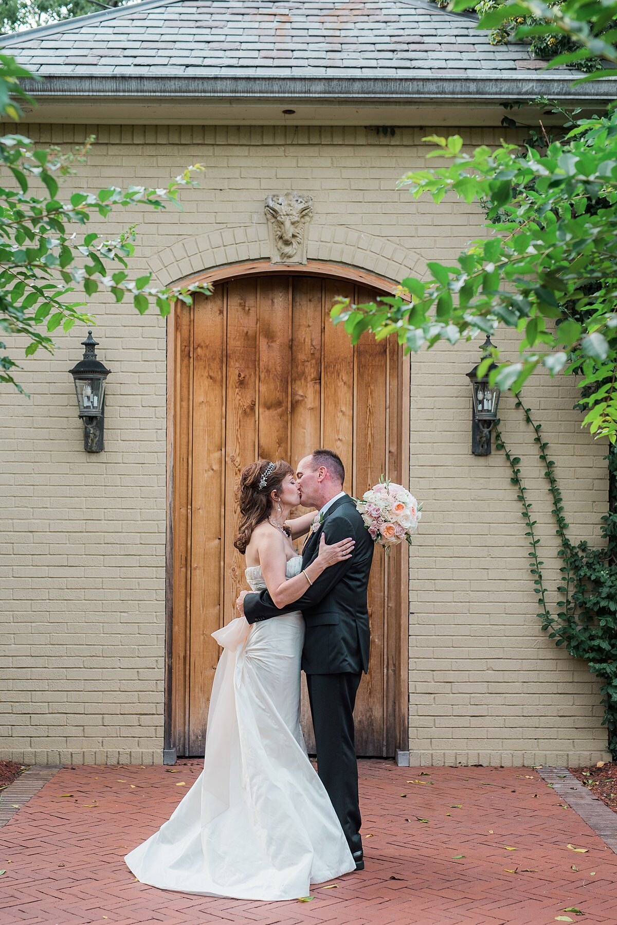 The bride, in a long satin gown and tiara kisses the groom in a dark gray suit at East Ivy Mansion while holding a bouquet of white and blush flowers