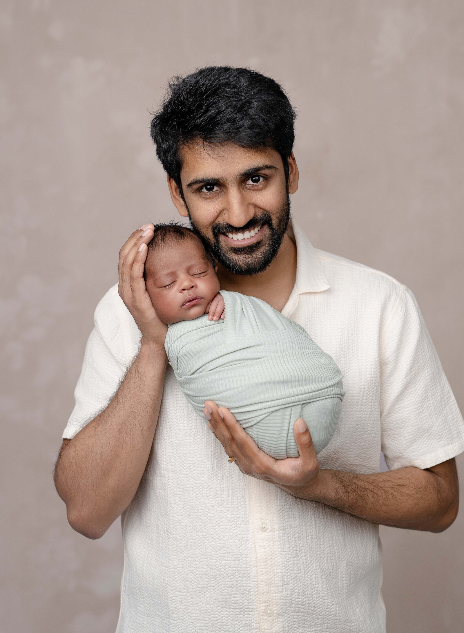 Dad is holding baby in sage swaddle and looking at camera. Newborn Photography session photographed at Jenni Nikki's photo studio in Woodstock, GA, near Atlanta.