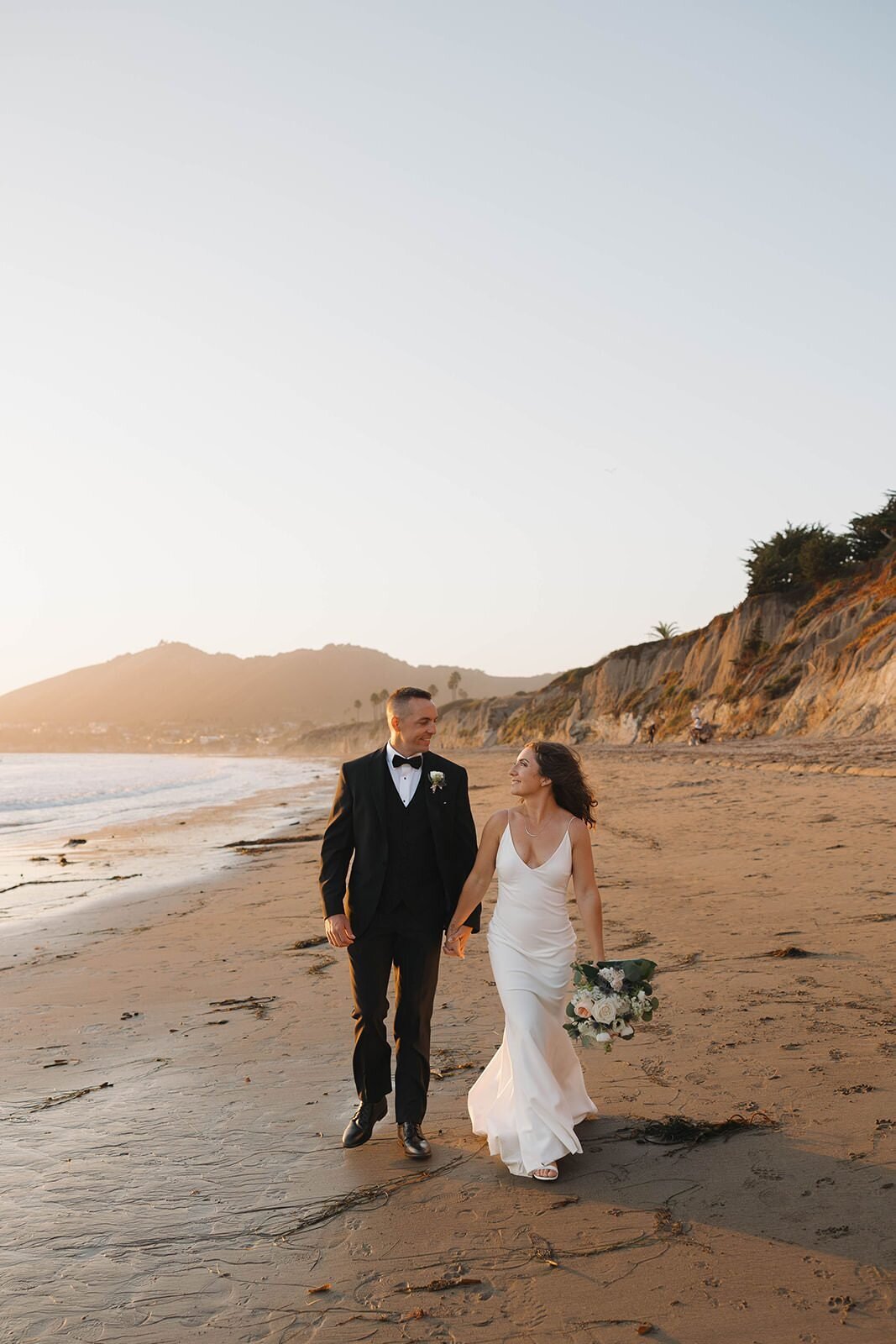 Bride and groom walking on the sand below the cliffs in Pismo Beach