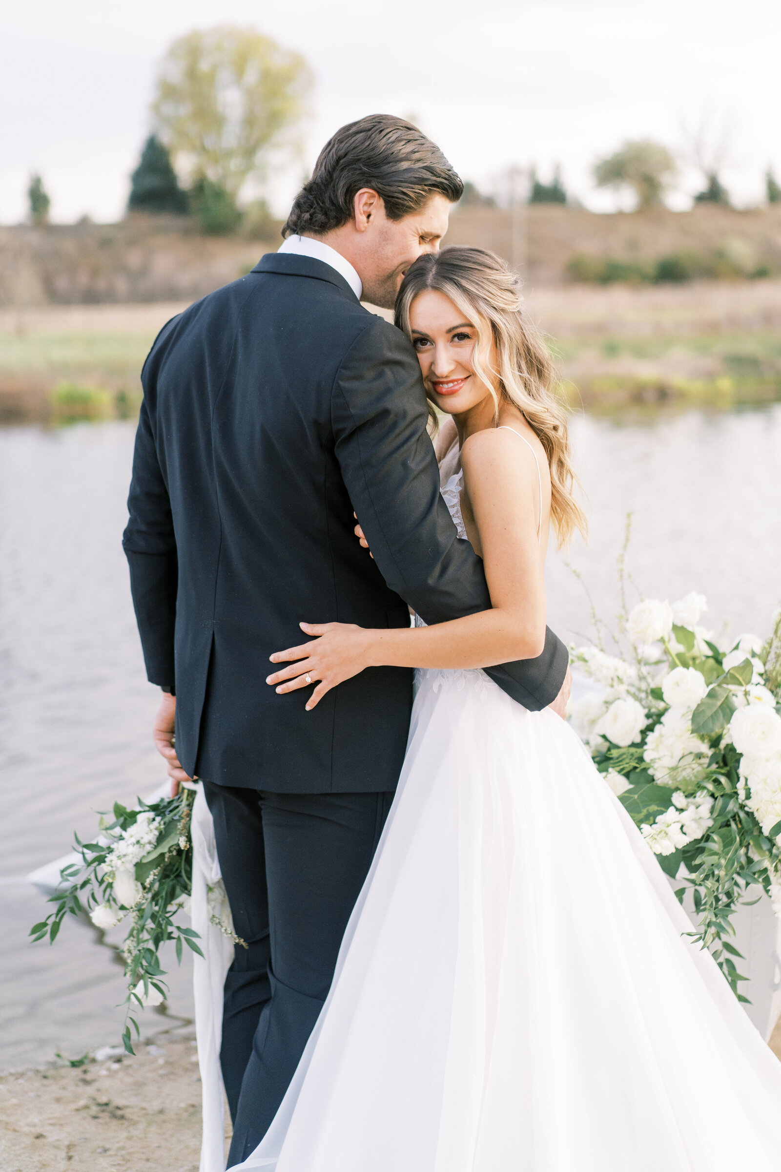 Portrait of a bride and groom in a white wedding gown and black tuxedo, holding each other and a bouquet in front of a lake